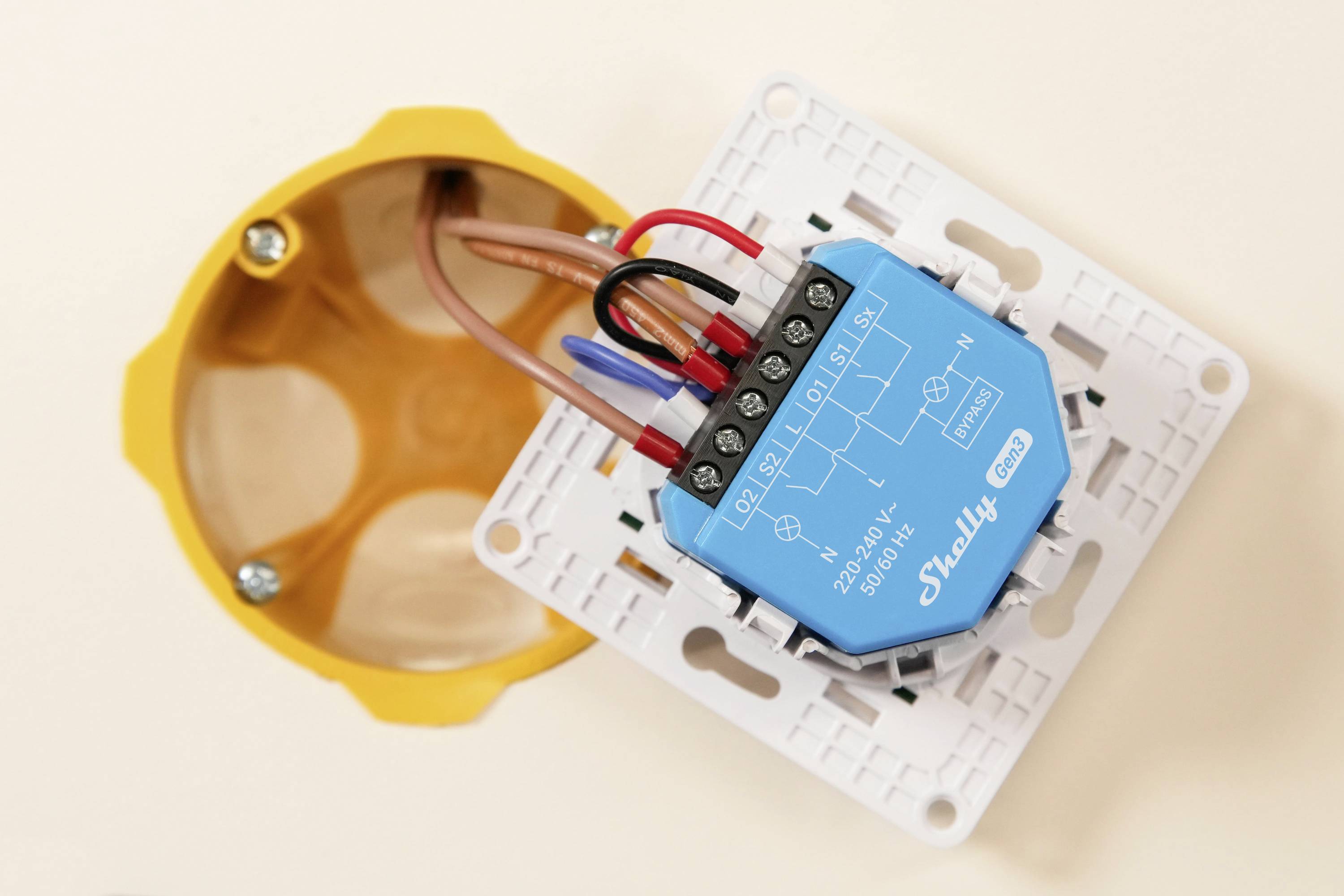 Egy kék Shelly intelligens kapcsoló látható bekötve és rögzítve egy fehér elektromos elosztódobozban, felkészülve a sárga fali aljzatba történő beépítésre.