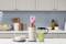 Egy NutriBullet turmixgép áll a konyhapulton, róaszínű smoothie hozzávalókkal és diófélékkel tele. Mellette egy pohár zöld smoothie van.
