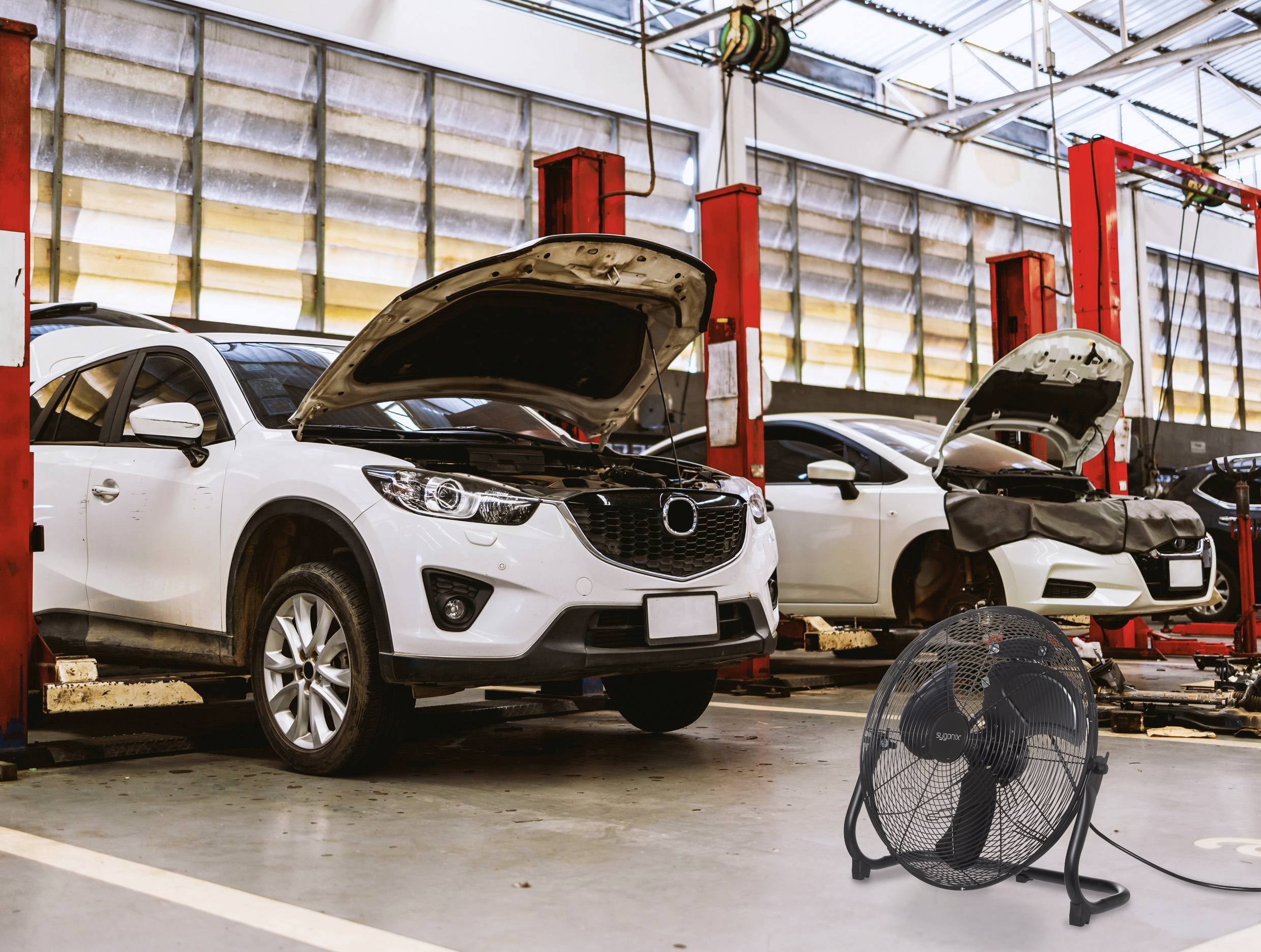 Két autó nyitott motorháztetővel hidraulikus emelőkön egy autószerelő műhelyben; egy nagy ventilátor van az előtérben.