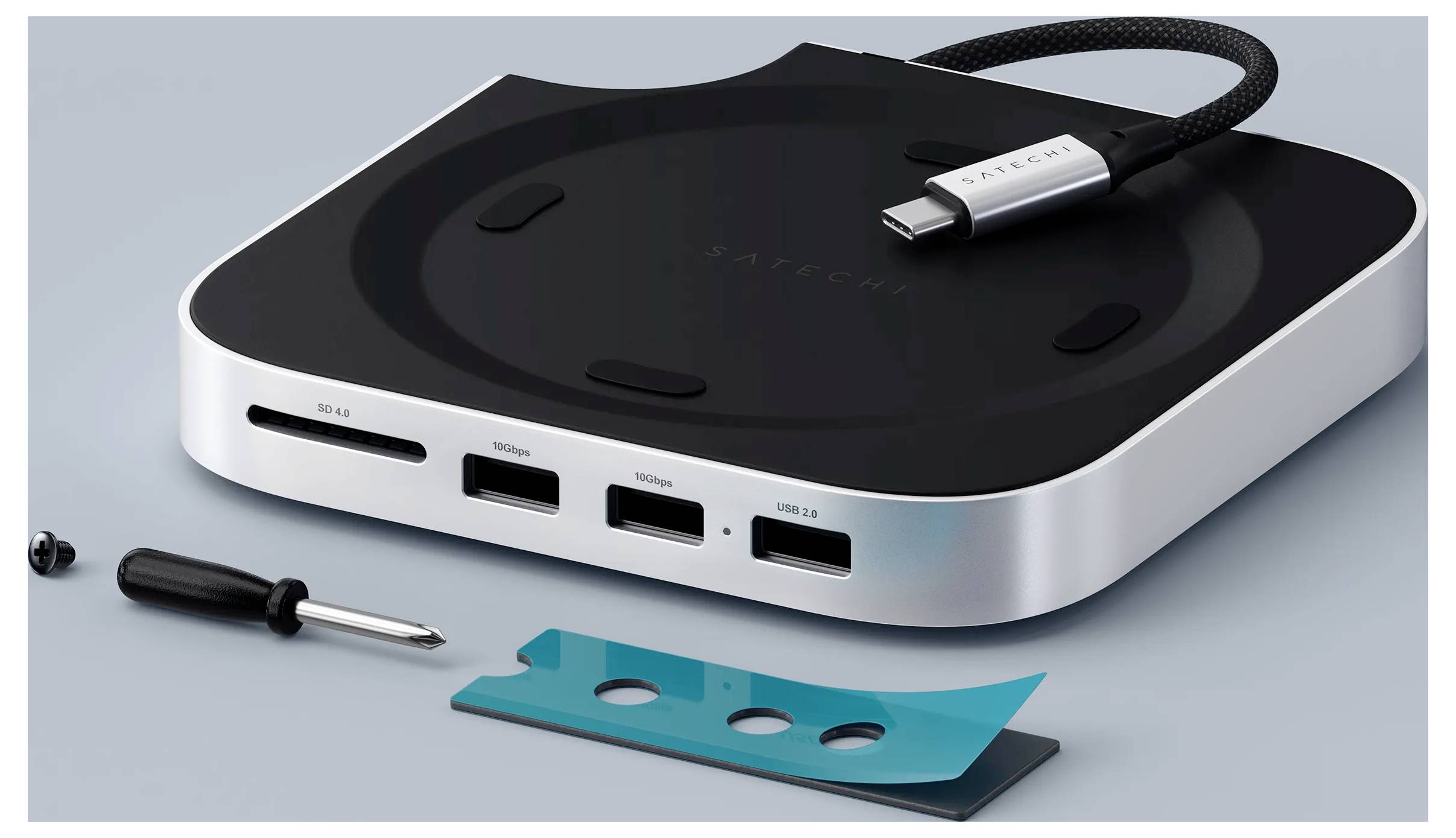 Egy karcsú USB-C elosztó több HDMI és USB csatlakozóval, egy csavarhúzóval és egy zöldeskék csíkkal egy szürke felületen.