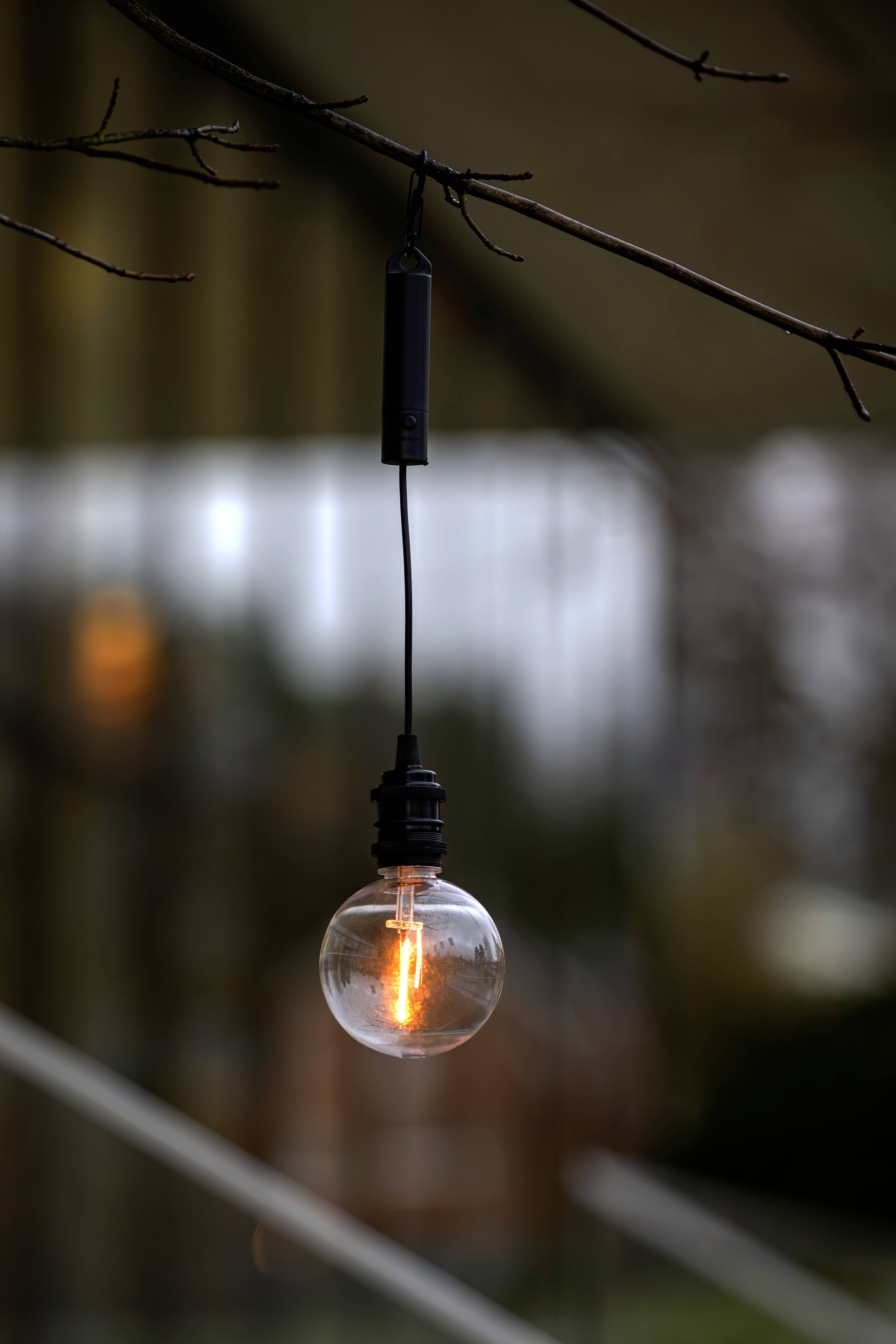 Egy felakasztott Edison-stílusú villanykörte lóg egy faágról a szabadban, meleg, körben sugárzó fényt árasztva egy elmosódott épület háttere előtt.