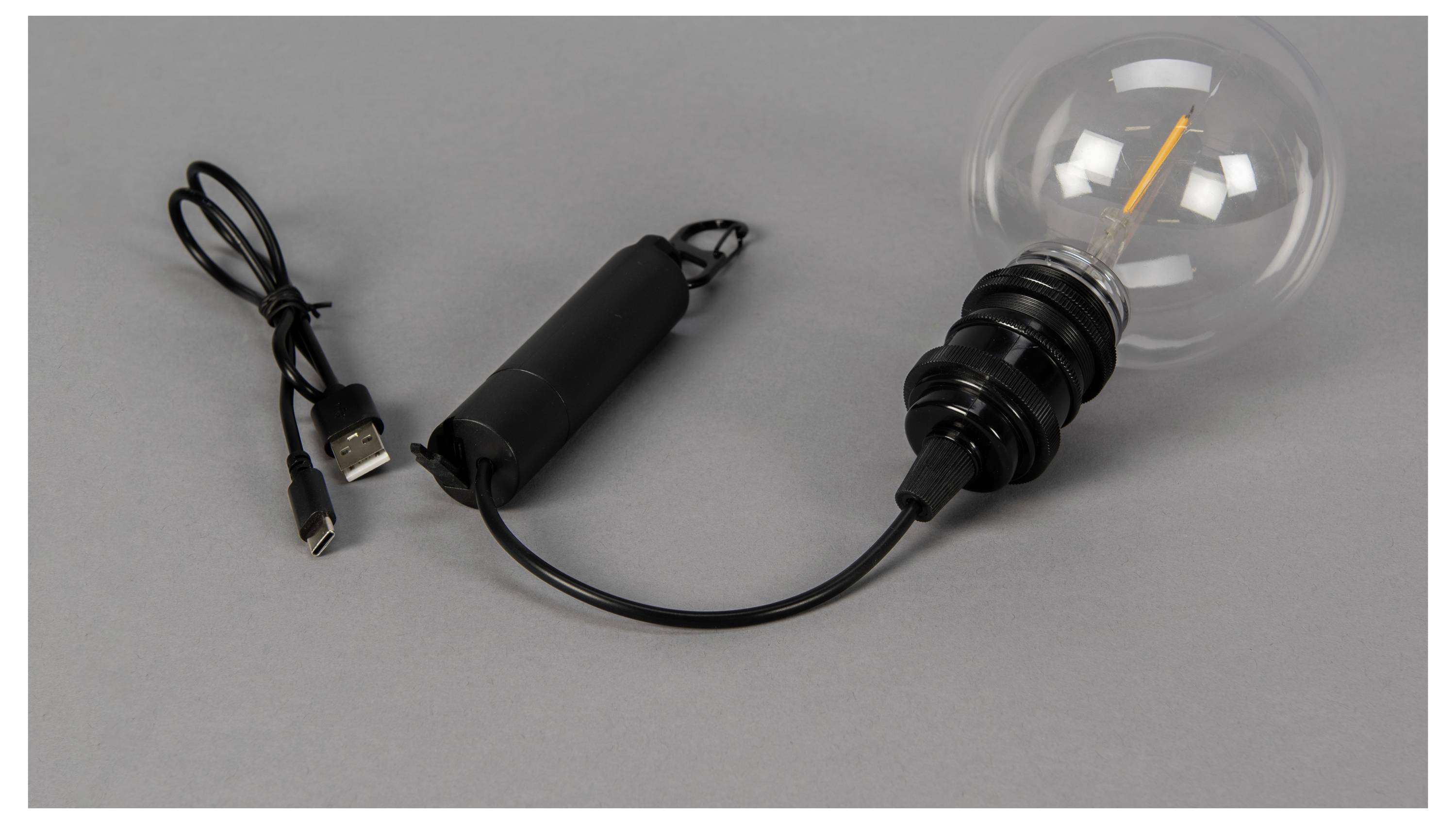 Egy USB-táplálású izzólámpa, amelynek fényforrása egy szálizzó, fekete zsinórja és hengeres tápegysége van, szürke felületen fekszik.