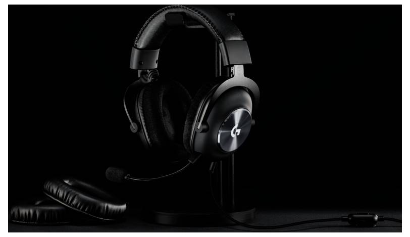 Egy fekete gaming headset fémlogóval és mikrofonnal egy állványon fekszik, körülötte extra fülpárnák, sötét háttér előtt.