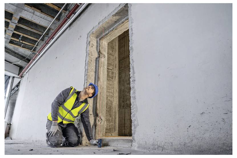 Egy sárga mellényt és kék védősisakot viselő építőmunkás egy félig kész épület ajtókeretét vizsgálja, ellenőrizve annak egyenességét és stabilitását.