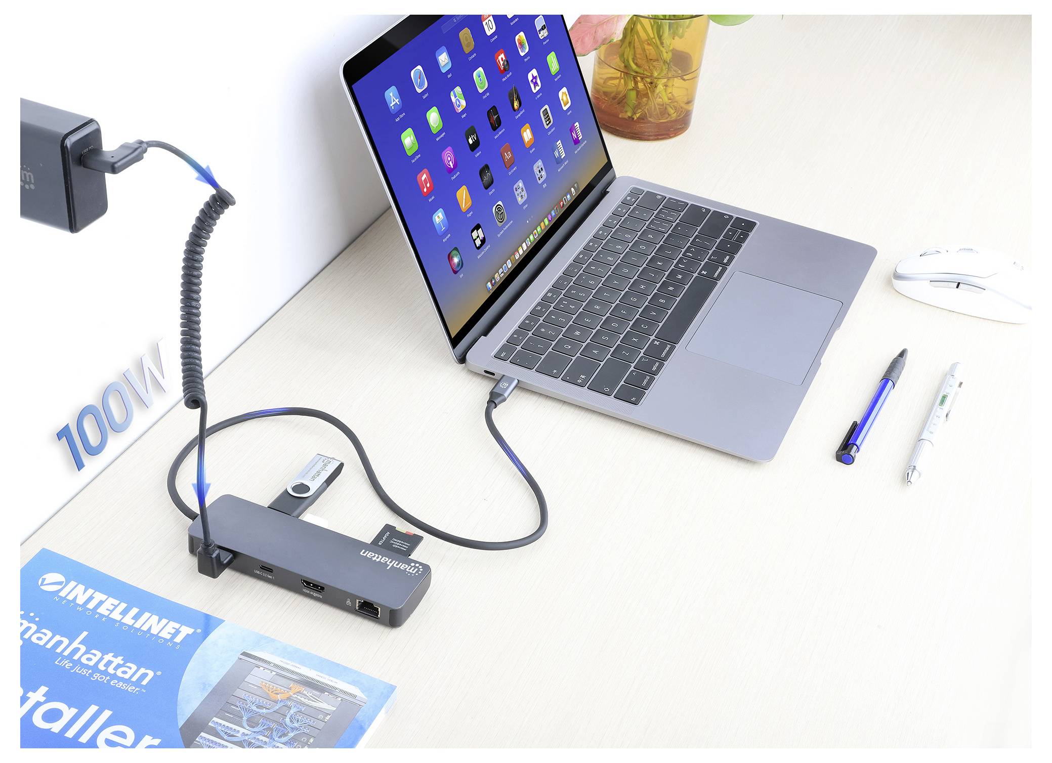 Egy laptop dokkolóállomáshoz csatlakoztatva, különböző kábelek segítségével, egy egér, tollak és egy füzet mellett egy íróasztalon. A hálózati adapter 100W-tal van felcímkézve.