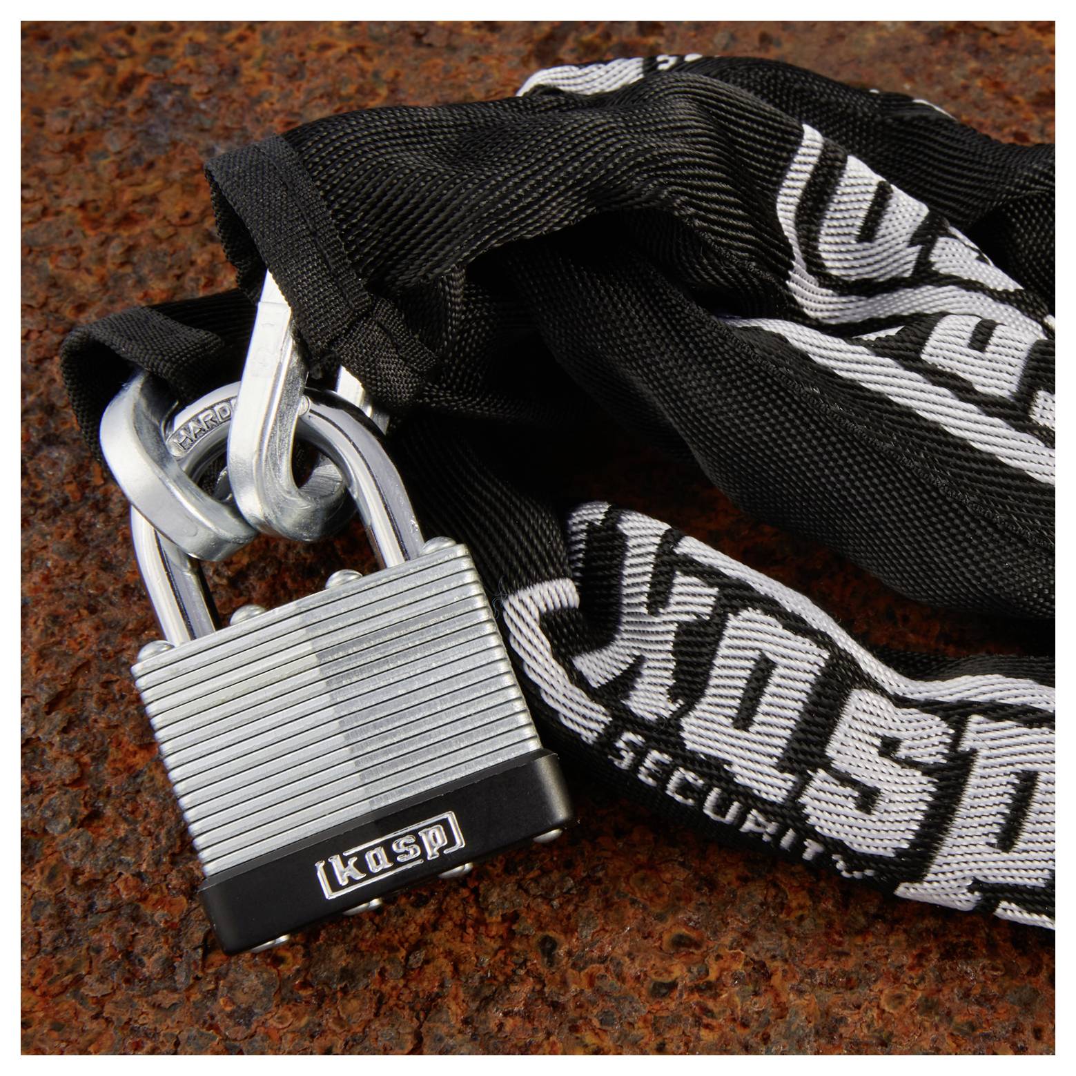 Egy masszív fémlakattal egy fekete lánc van lezárva, amelyen a 'Kasp Security' felirat olvasható egy rozsdás fémfelületen, ami erős védelmet sugall a lopás ellen.
