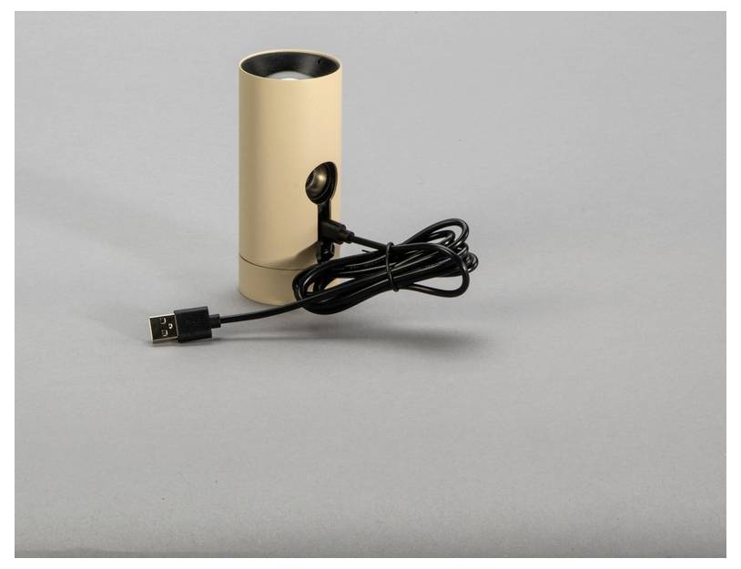 Egy bézs színű, hengeres alakú hangszóró USB-kábellel együtt fekszik egy szürke felületen.