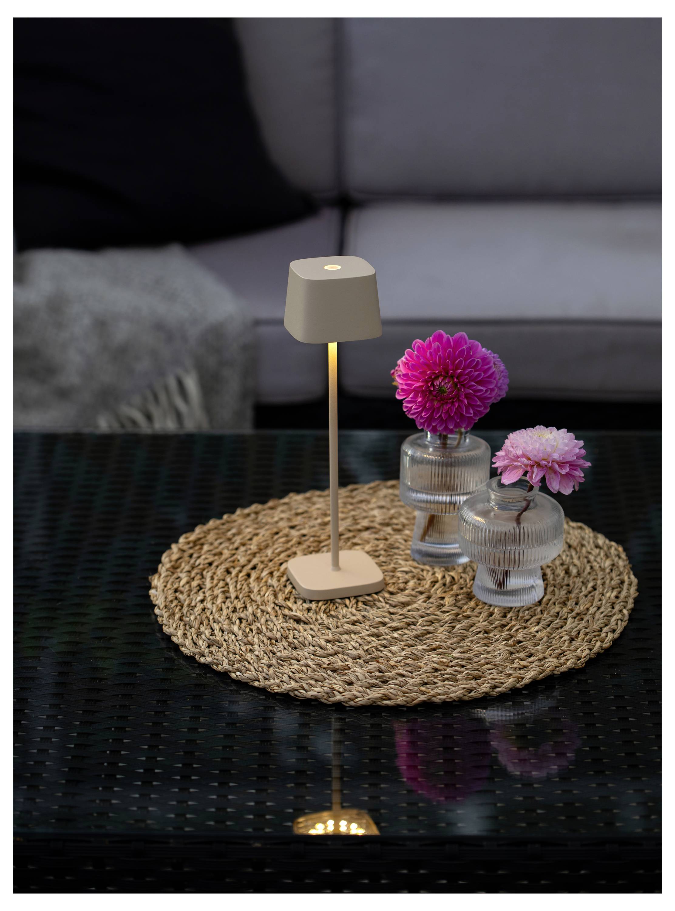 Egy modern asztali lámpa és két rózsaszín virág üvegvázákban egy szőtt szőnyegen helyezkedik el egy üveg dohányzóasztalon, a háttérben egy kanapéval.