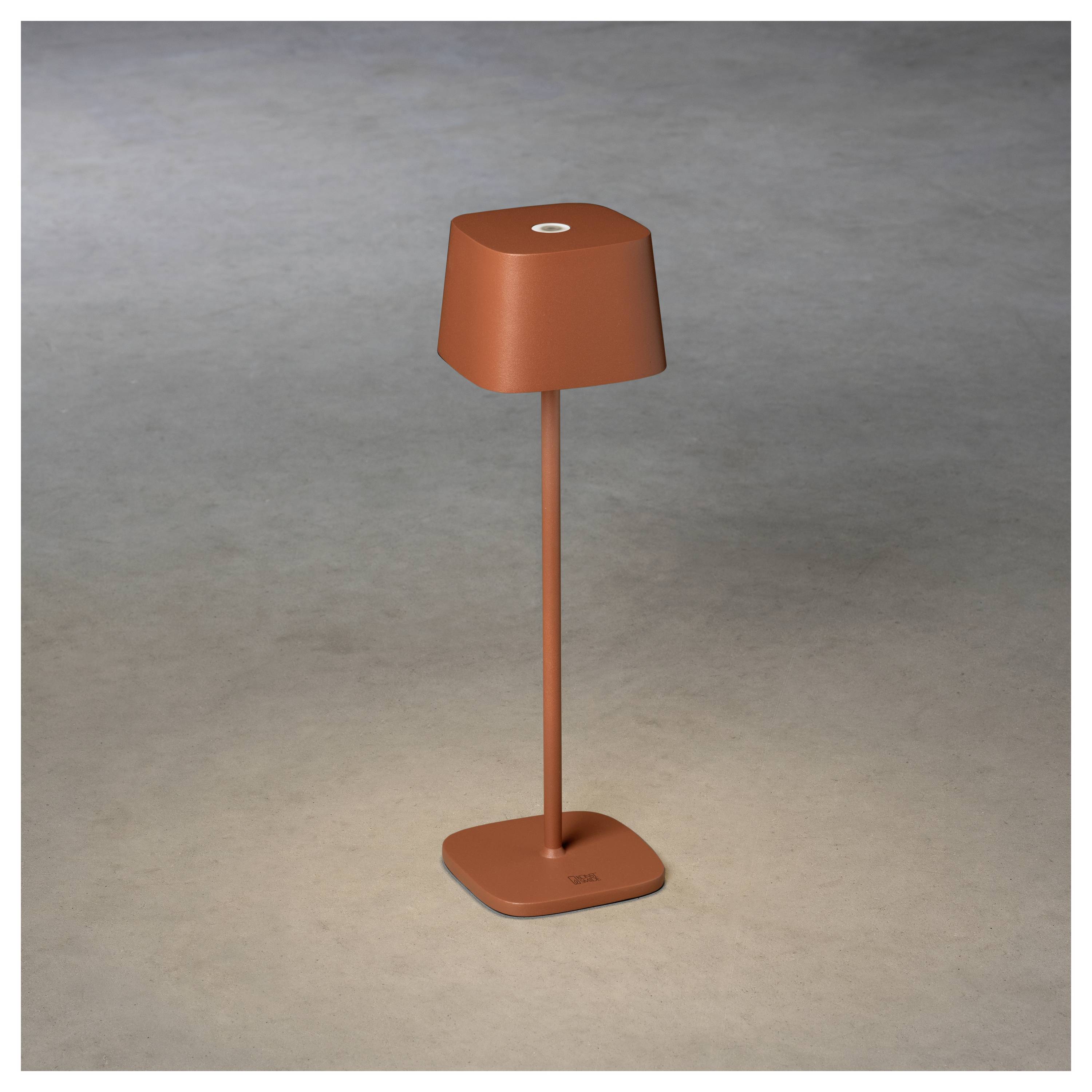 Egy minimalista vörös asztali lámpa négyzet alakú talapzattal és búrával, amely lágy fényt áraszt semleges háttér előtt.