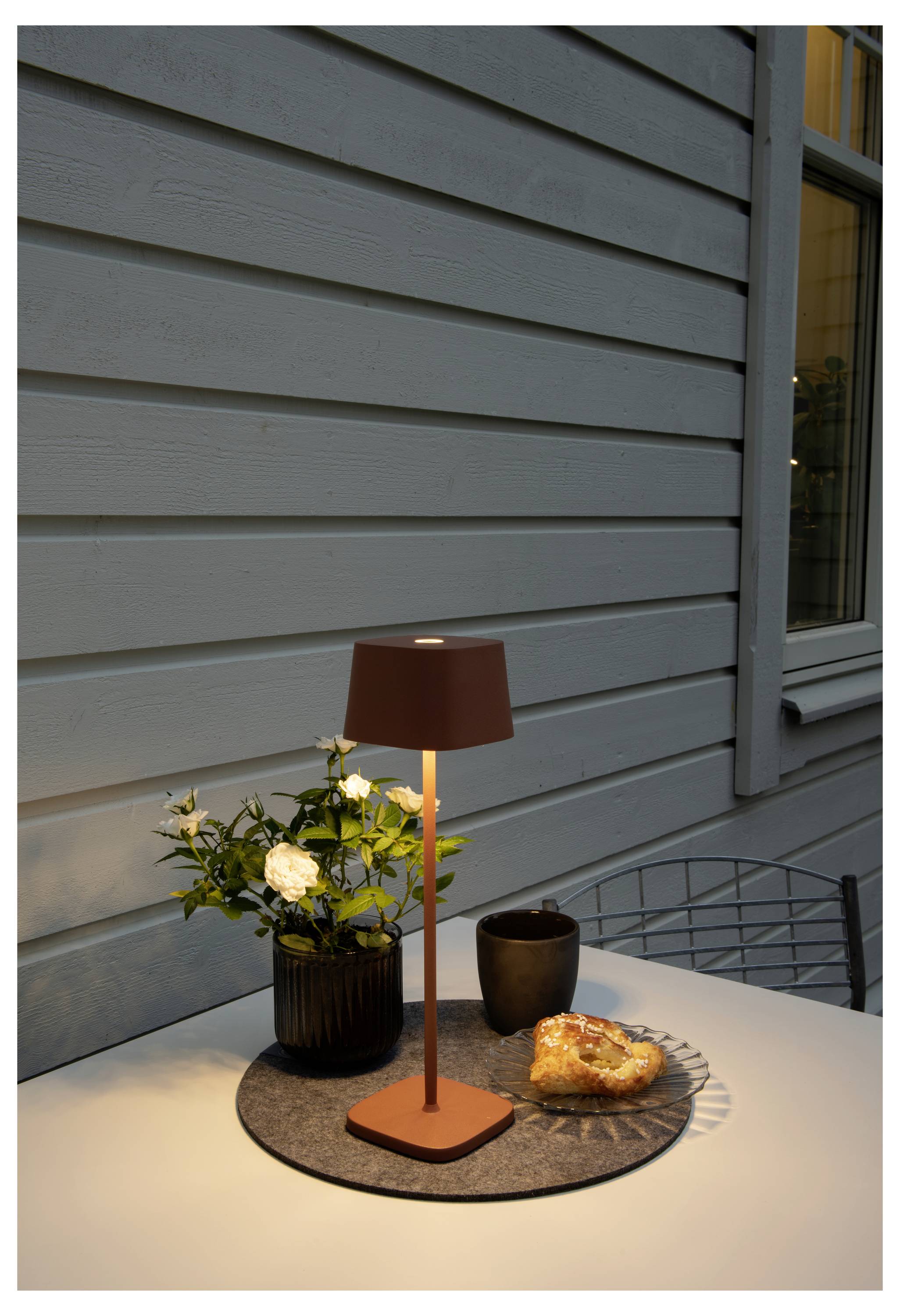 Egy kerek asztal áll egy fa fal előtt a szabadban, rajta egy kisméretű lámpa, egy cserépben lévő növény, egy kávéscsésze és egy sütemény.