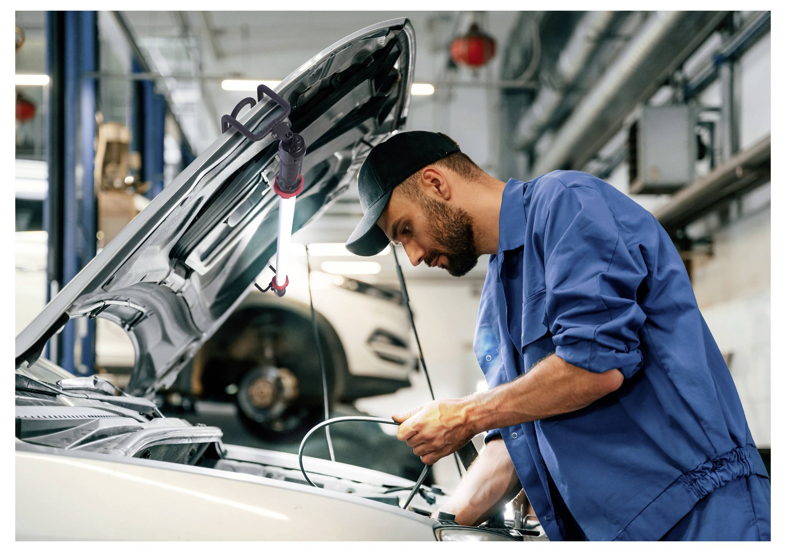 Egy kék uniformisban dolgozó szerelő egy autó motorján dolgozik a garázsban, a motorháztető nyitva van és szerszámok vannak a kezében, intenzíven koncentrál a javításokra.