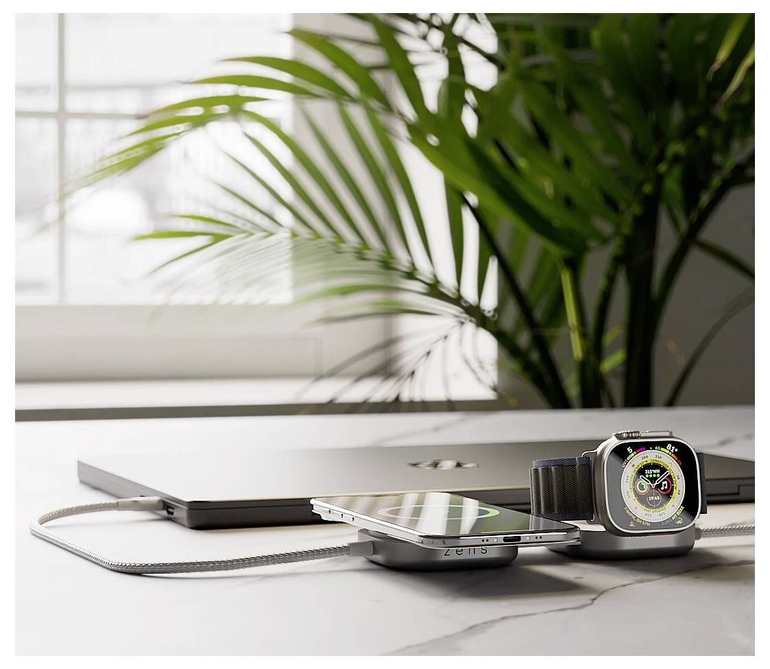 Egy okostelefon és egy okosóra tölt egy kettős vezeték nélküli töltőn, amelyet egy márvány asztalon helyeztek el egy zárt laptop és egy nagy növény mellett.