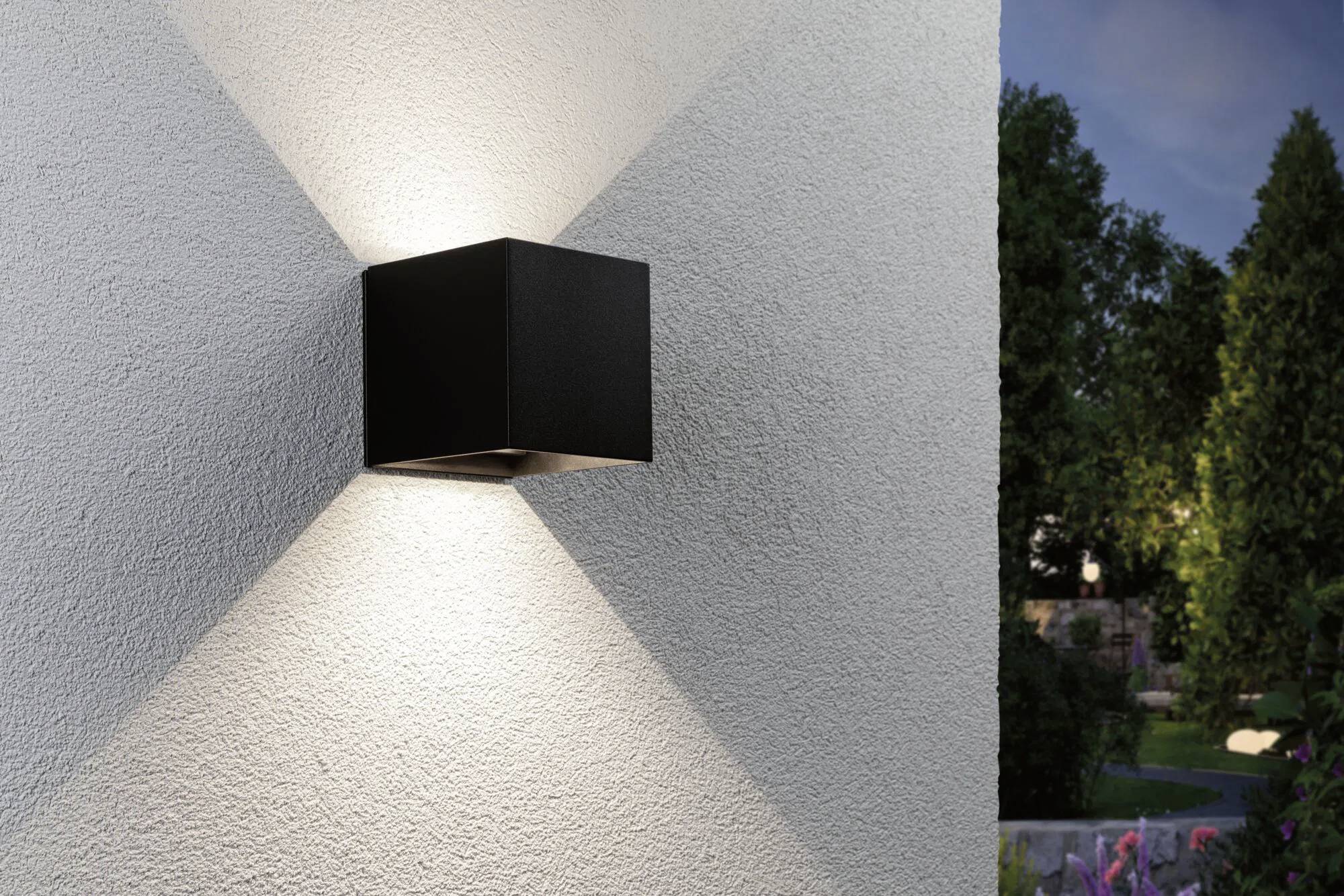 Egy modern fekete fali lámpatest egy strukturált fehér külső falon, szimmetrikus fénymintázatokat vetve. A háttérben kert és fák láthatók.