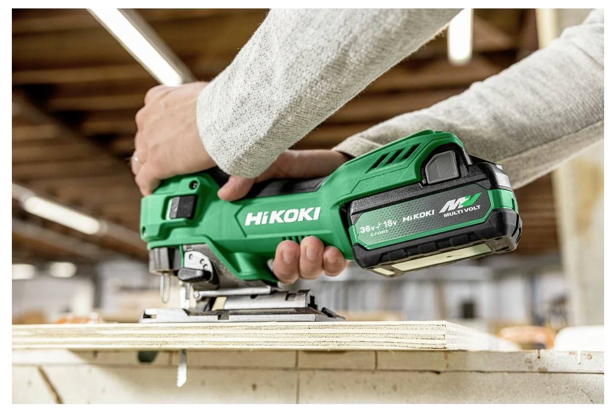 Egy személy egy zöld, akkumulátoros dekopírfűrészt használ egy fafelületen egy műhelyben, kiemelve a szerszám márkáját és akkumulátoros jellegét.