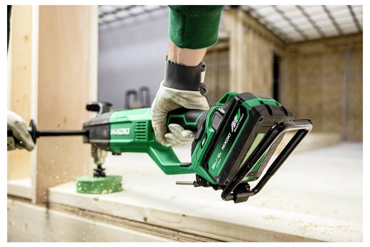 Egy ember egy Hikoki rezgőfűrésszel fadeszkát vág bent, kesztyűt viselve. A fűrészpor látható a dinamikus műhelykörnyezetben.