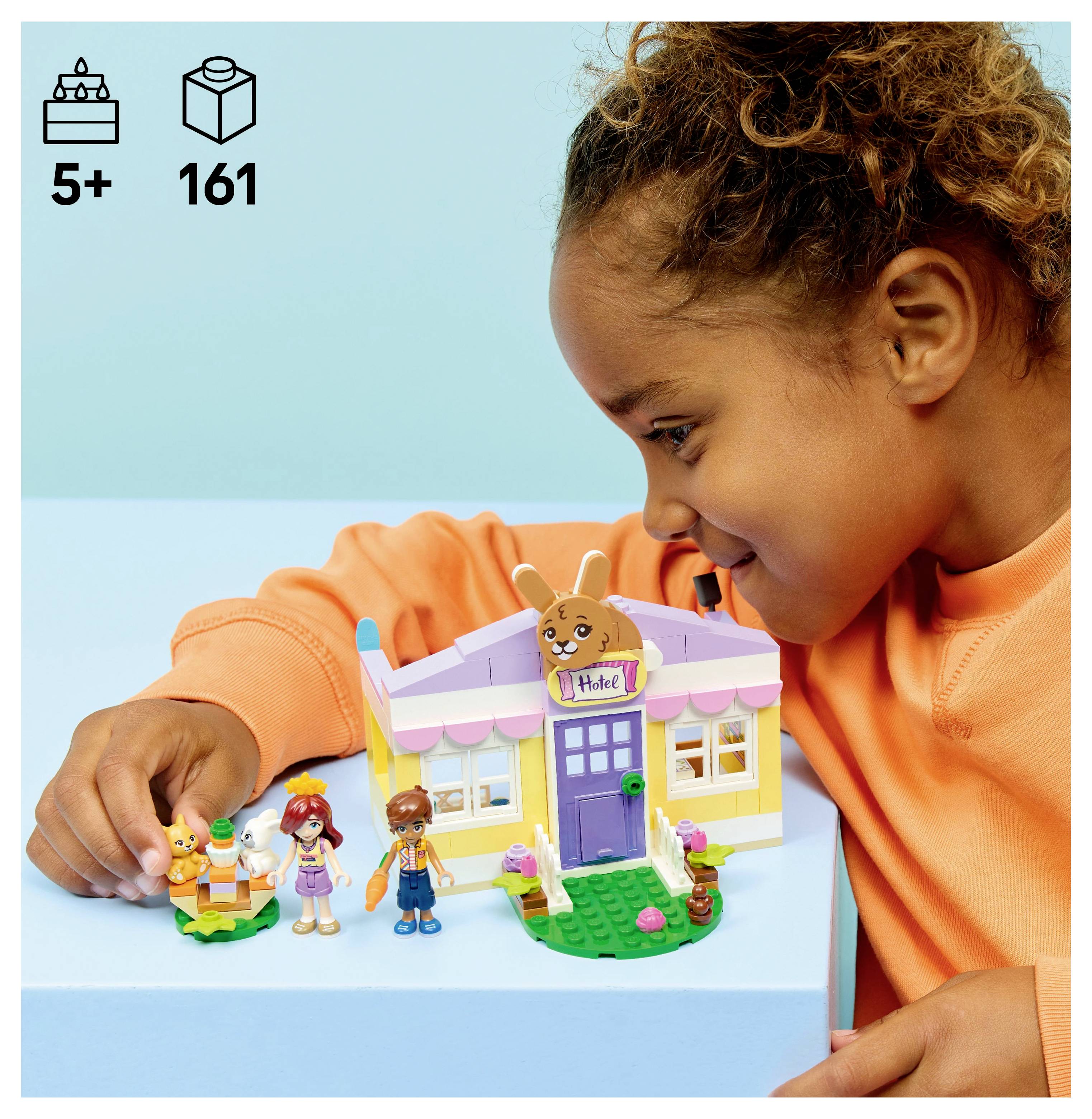 Gyermek mosolyog, miközben egy LEGO szett játszik, amely egy kis sárga házat és figurákat tartalmaz; a doboz jelzi a '5+' életkort és a '161' darabot.