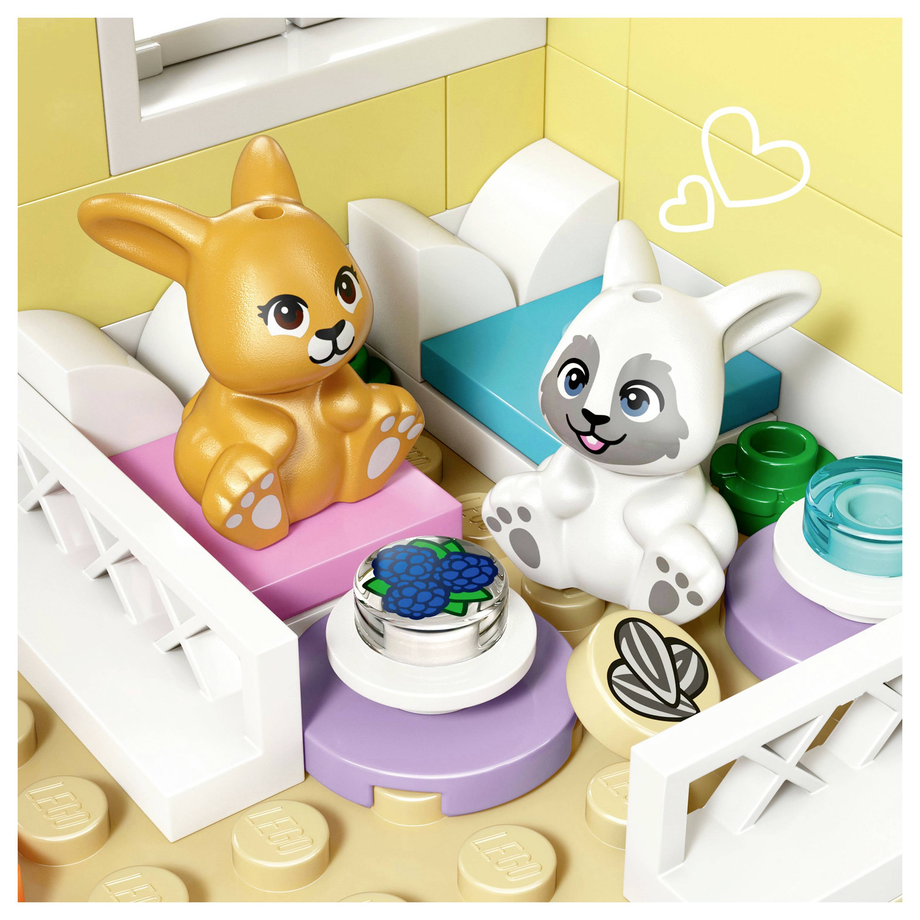 Két játék nyuszi ül színes ágyakon egy játszószobában. Egy kis asztal virágdísszel van köztük elhelyezve. Két szívszimbólum van fölöttük megrajzolva.