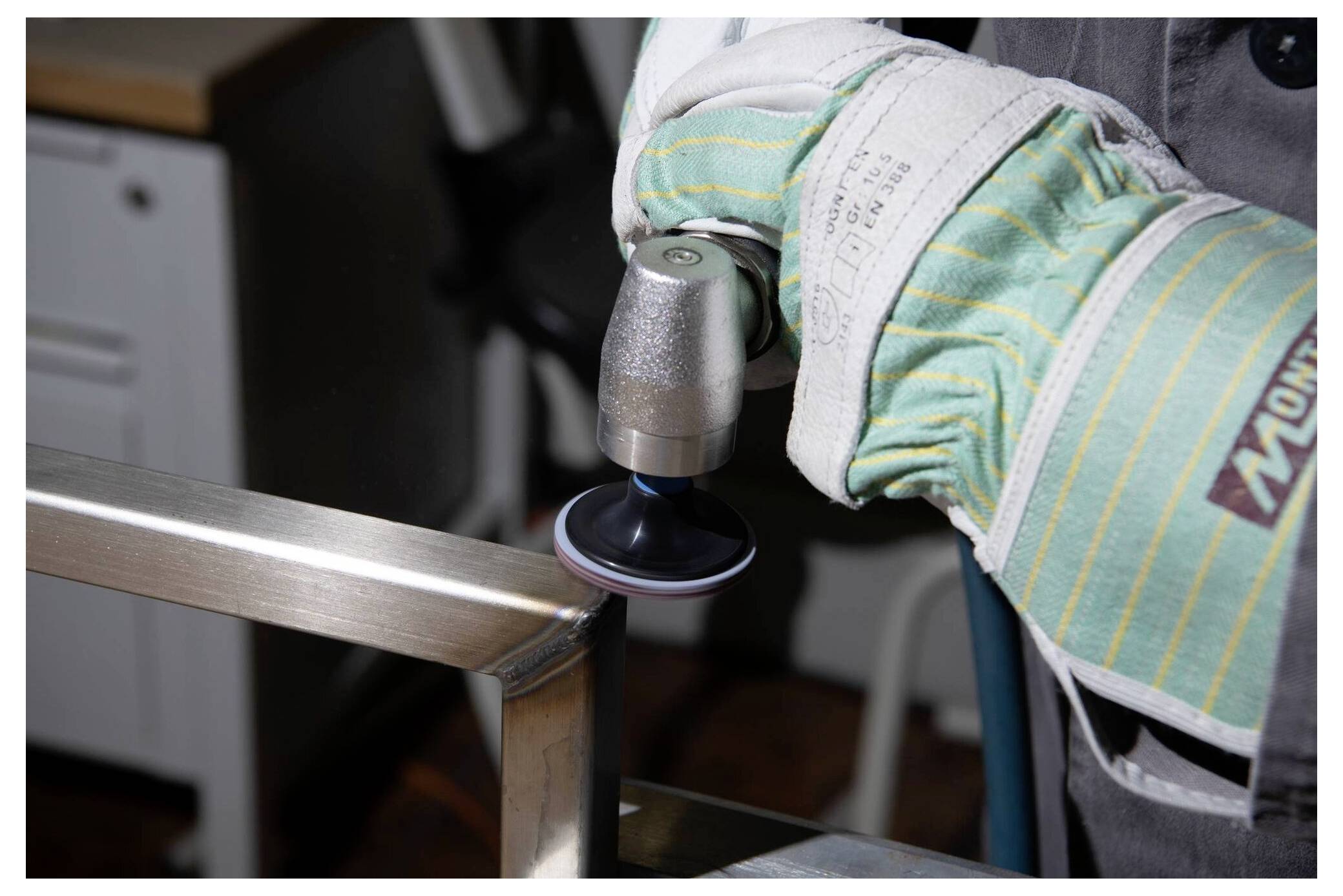 Közeli felvétel egy zöld csíkos kesztyűt viselő kézről, amint egy power tool-lal polírozza egy fémobjektum szélét egy műhelyszerű környezetben.