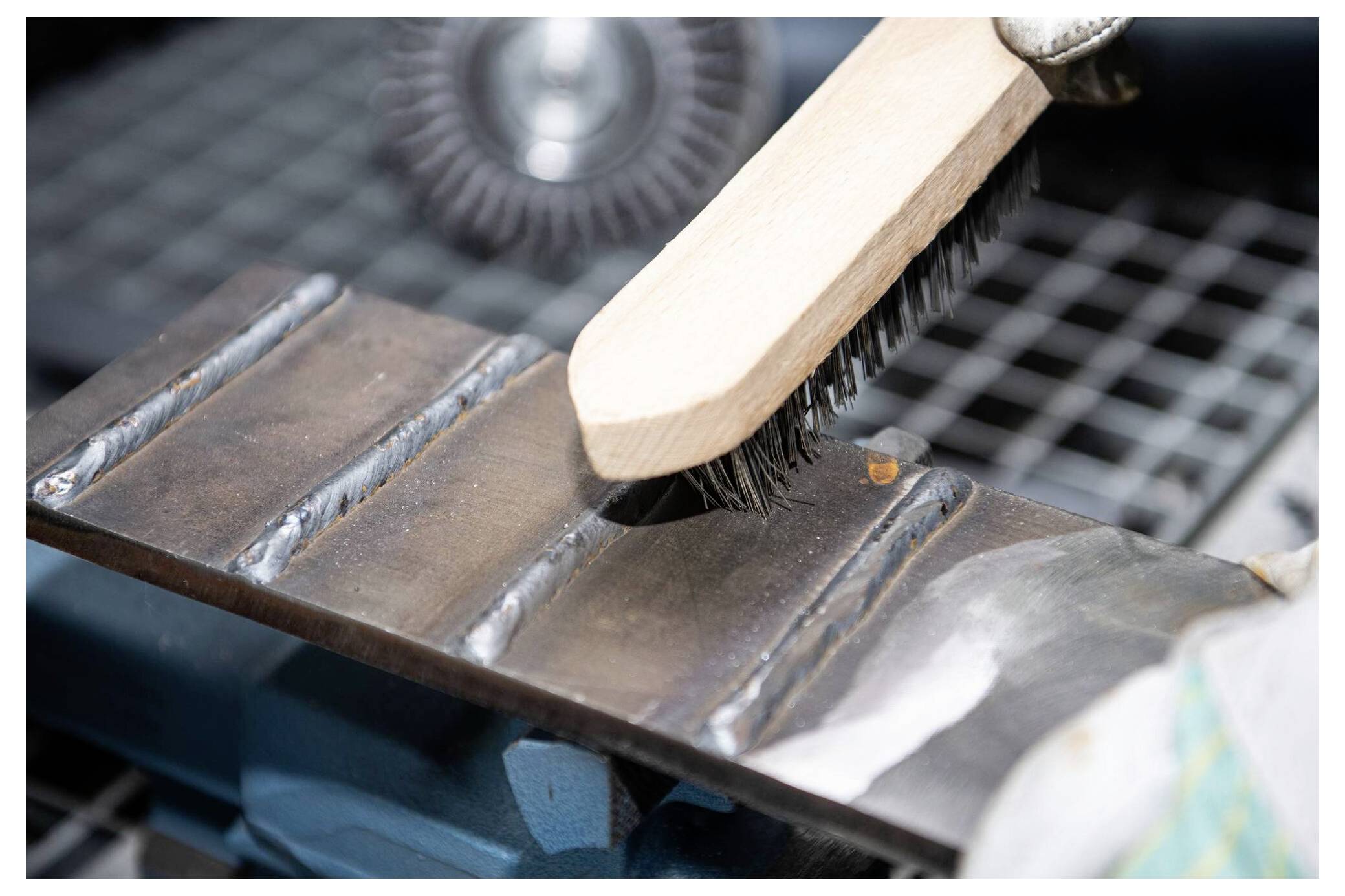 Egy drótecset egy féktárgy felületét tisztítja hegesztési varratokkal egy munkapadon, kiemelve a fémfelület-előkészítés folyamatát.