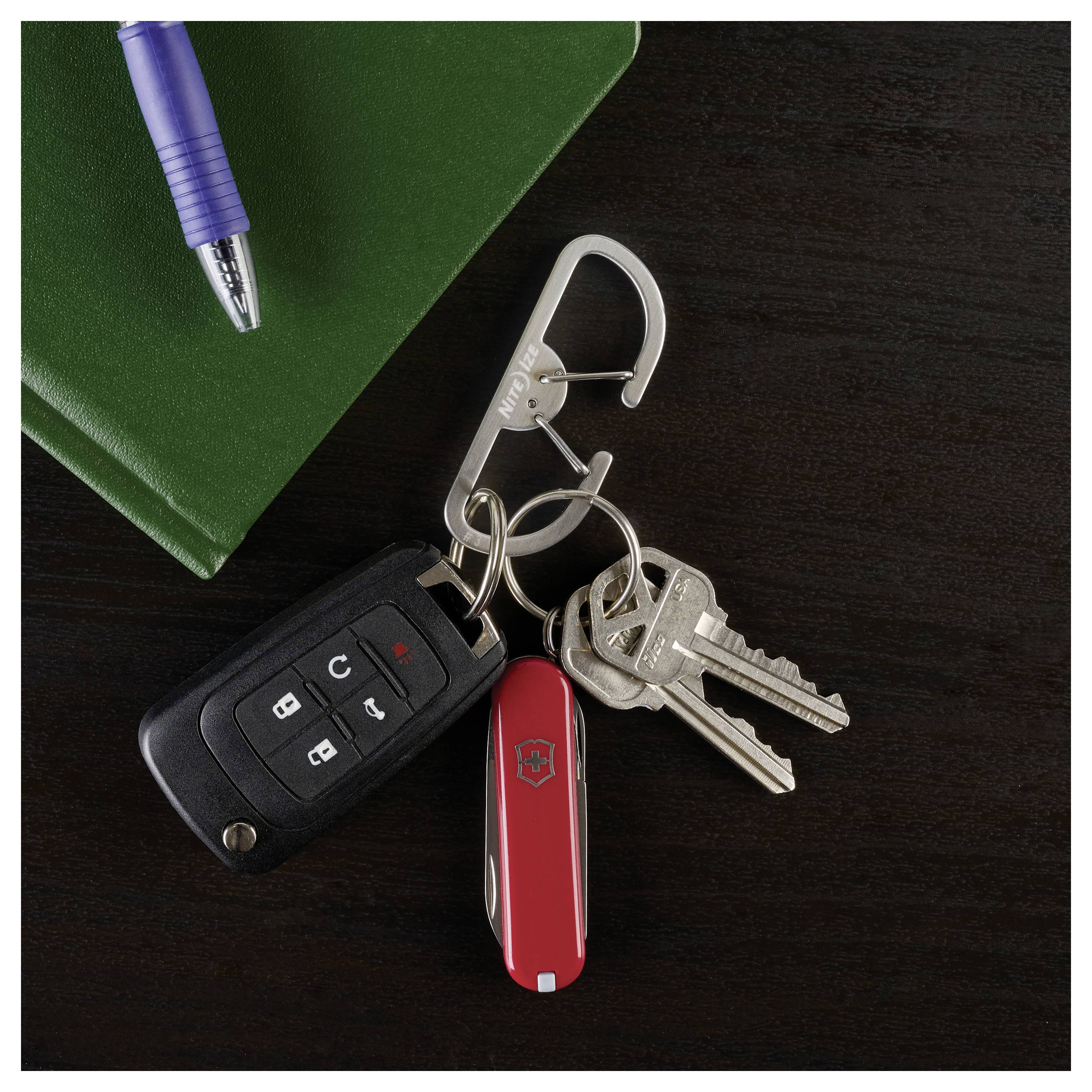 Egy piros svájci bicskával és egy autós kulcstartóval ellátott kulcsok egy ezüst karabinerhez vannak erősítve. Egy zöld füzet és egy kék toll van a közelben egy sötét felületen.