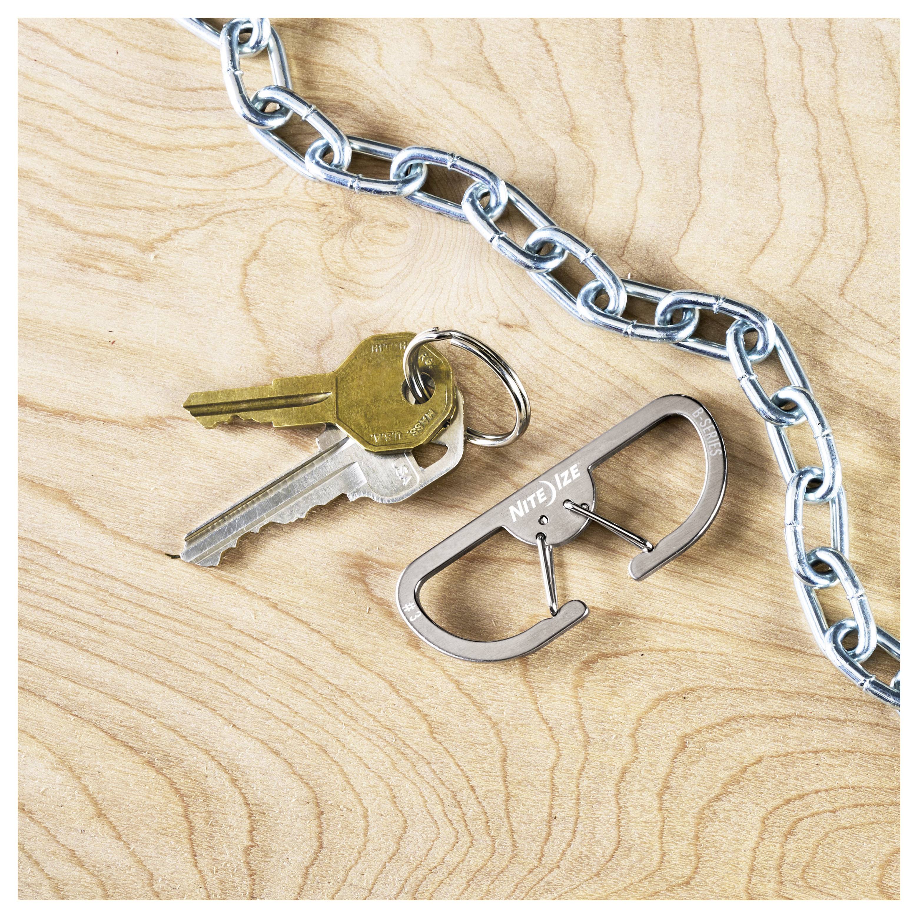 Egy kulcskészlet, amely két fém kulcsot és egy karabinerrel ellátott kulcstartót tartalmaz, egy fa felületen fekszik egy fém lánc mellett.