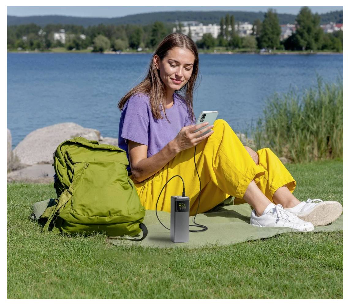 Egy nő füves területen ül egy tó partján, miközben egy hordozható napelemes töltőhöz csatlakoztatott okostelefont használ. Egy zöld hátizsák hever mellette.