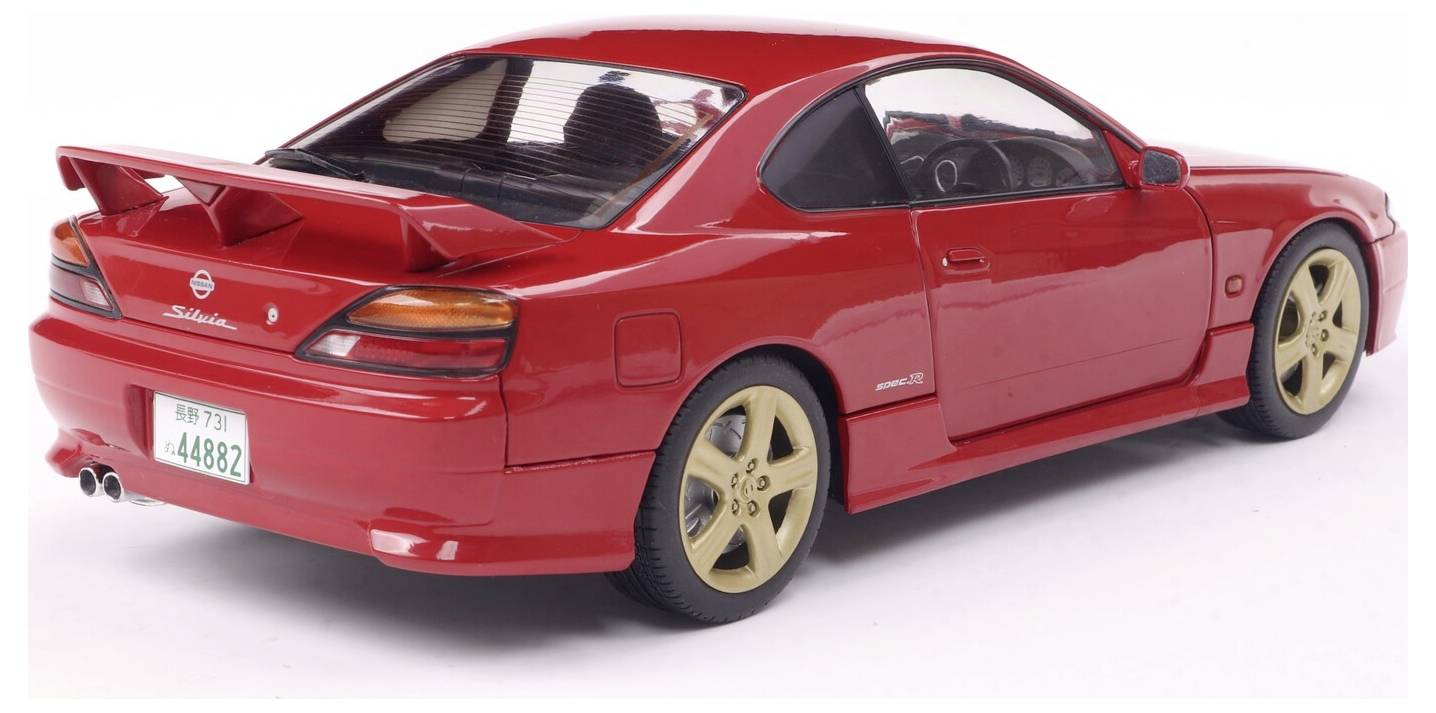 Egy piros sportautó hátsó spoilerrel és arany kerekekkel, hátulról jobbról nézve, fehér felületen parkolva.