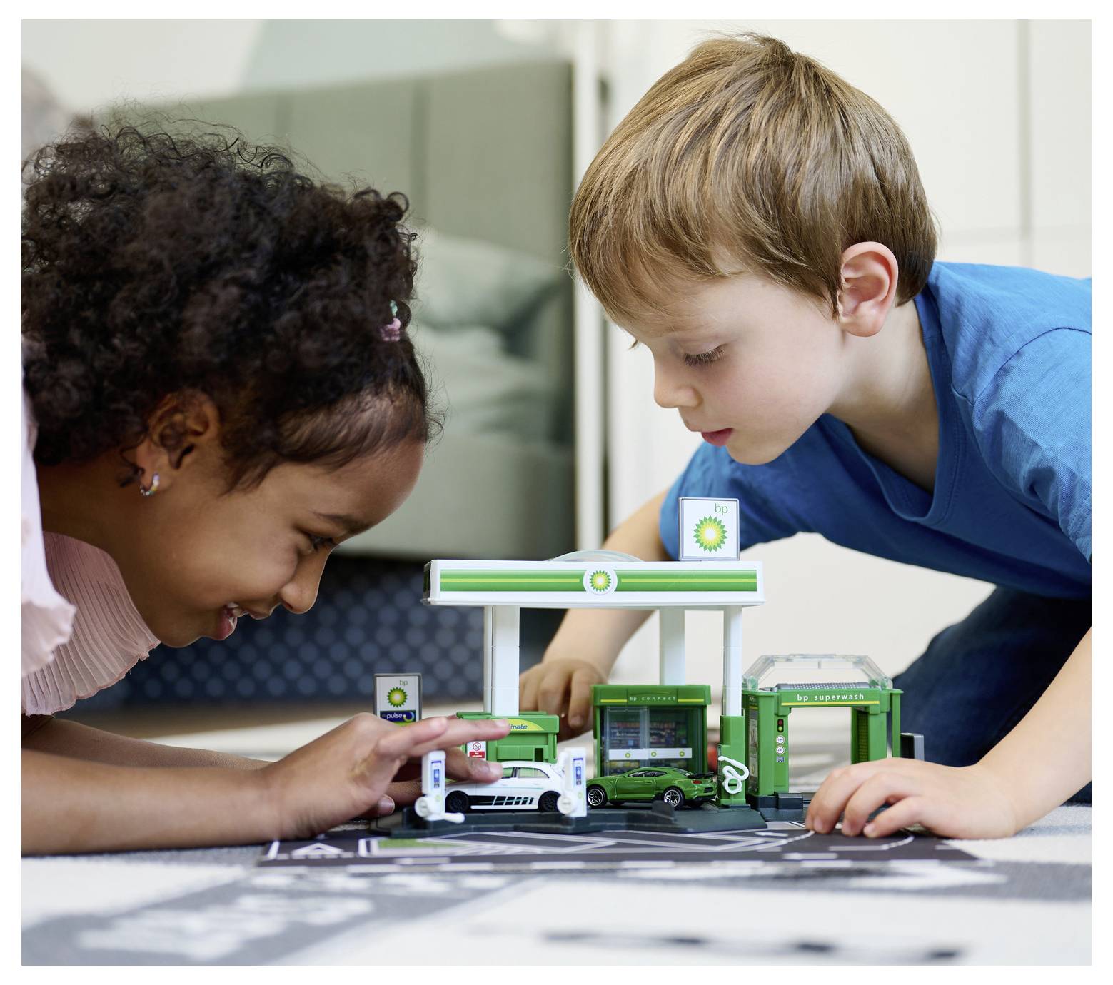 Két gyerek lelkesen játszik egy BP benzinkút játékszettel, figyelmesen tanulmányozva a miniatűr járműveket és a benzinkút apró részleteit.