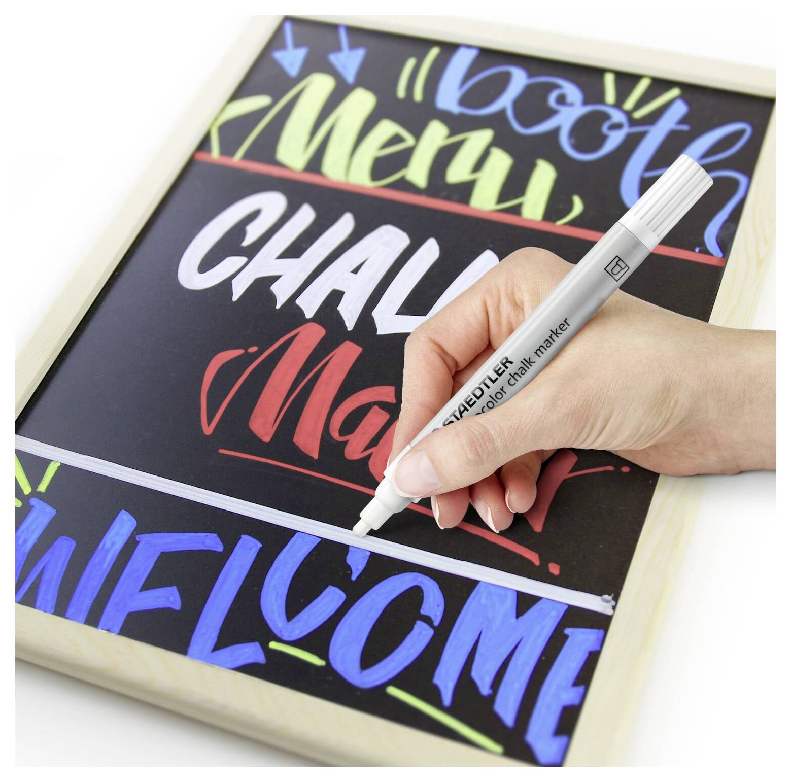 Egy kéz fehér krétamarkerrel színes szavakat ír egy táblára: 'Menü', 'Kihívás' és 'Üdvözlet'.