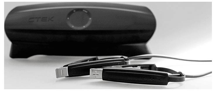 Egy fekete CTEK akkumulátortöltő a hozzá tartozó csipeszekkel fehér felületen fekszik, kiemelve az elegáns dizájnt és a praktikus funkcionalitást a járművek töltéséhez.