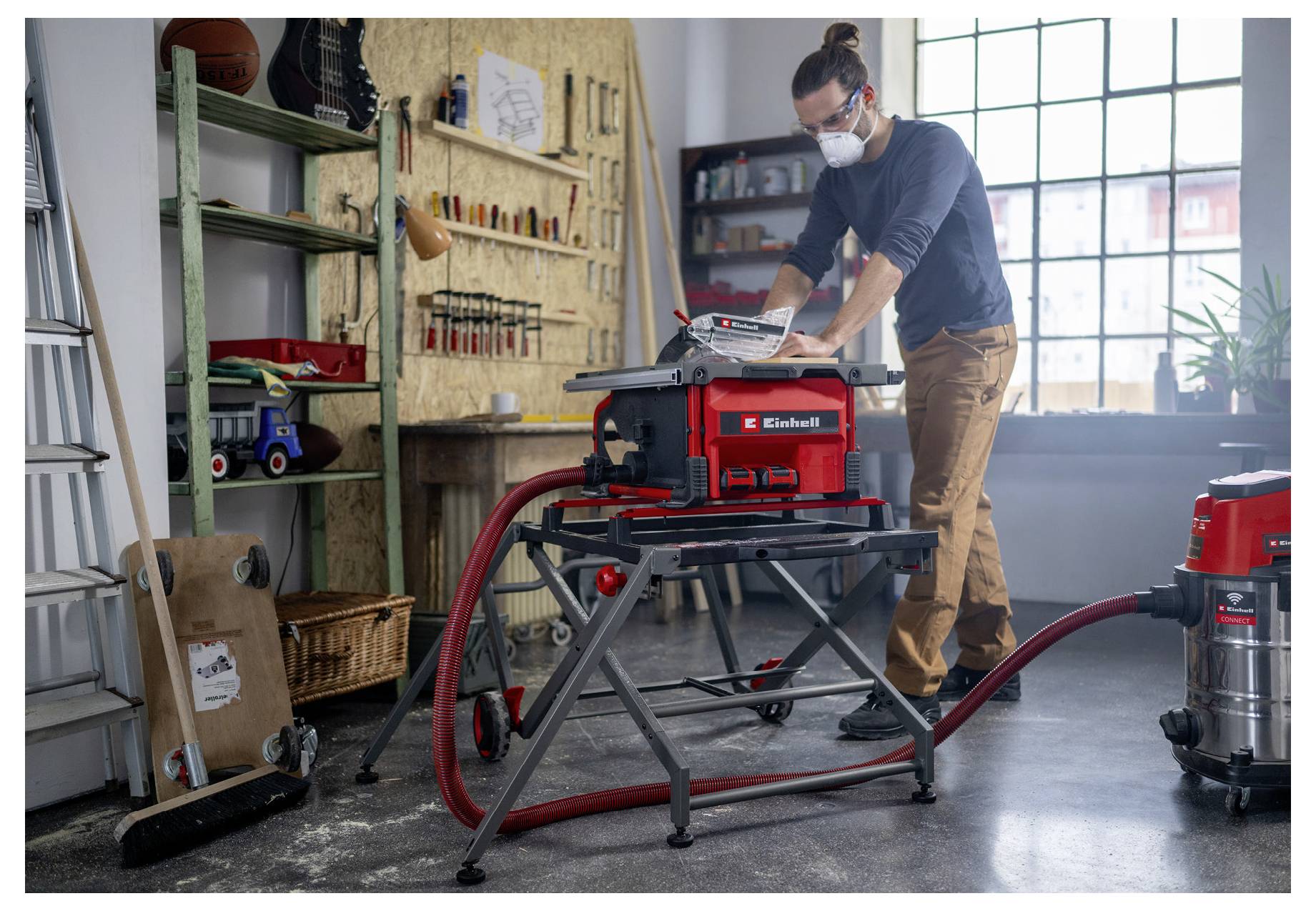 Egy ember egy műhelyben körfűrésszel dolgozik, védőfelszerelést viselve. Szerszámok és eszközök veszik körül, miközben nagy ablakon át napfény árasztja el a helyiséget.
