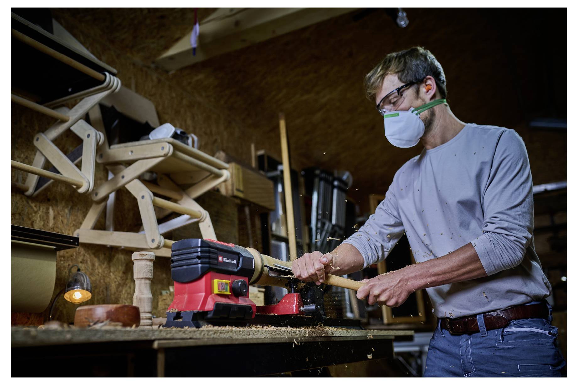 Egy maszkot viselő személy egyesztergapadon faanyagot megmunkál egy műhelyben. Szerszámok és fa szerkezetek láthatók a háttérben.