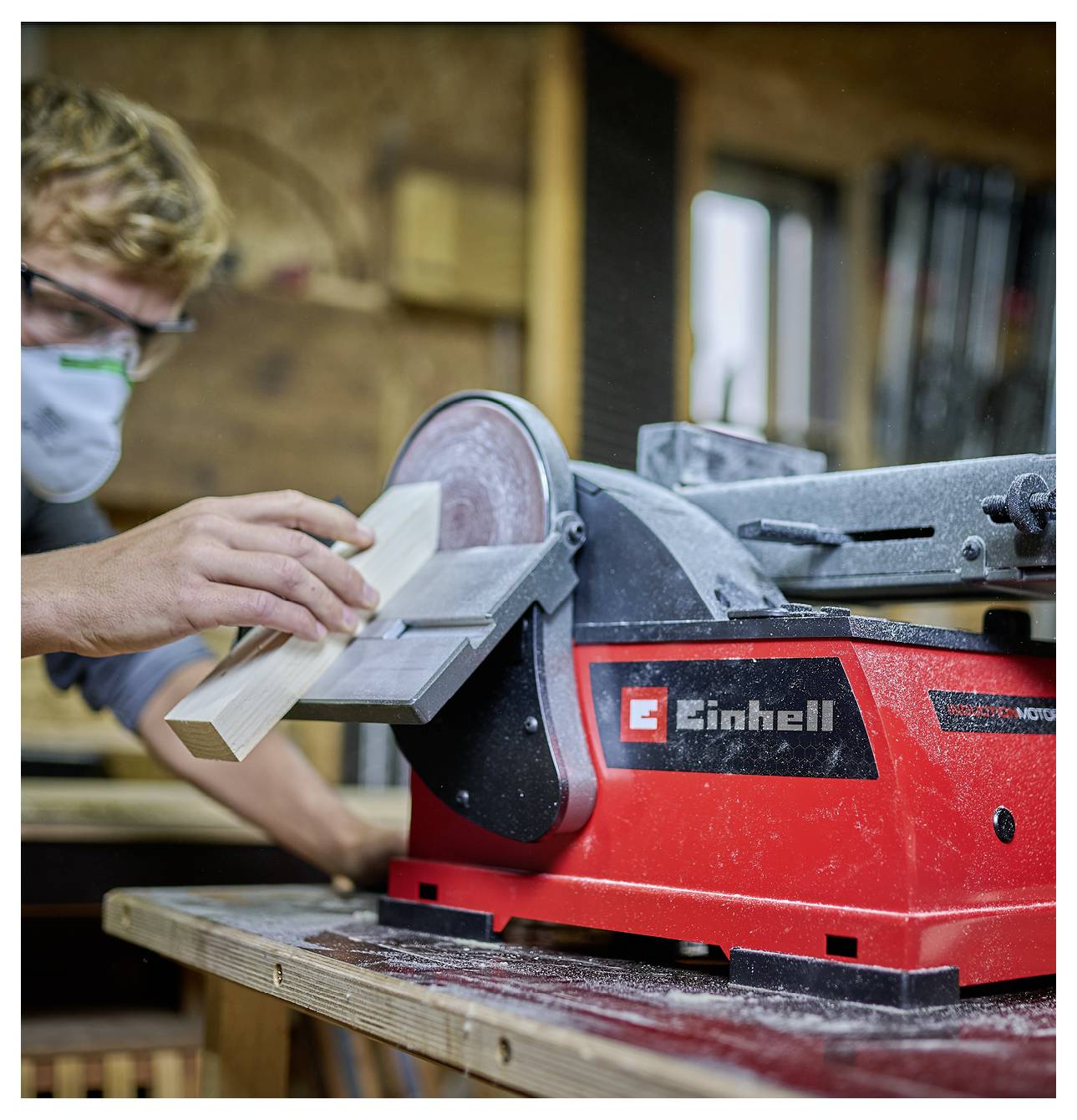 Egy személy, aki védőszemüveget és maszkot visel, egy piros elektromos padállványos köszörűgépet használ egy fadarab csiszolásához egy műhelyhelyiségben.