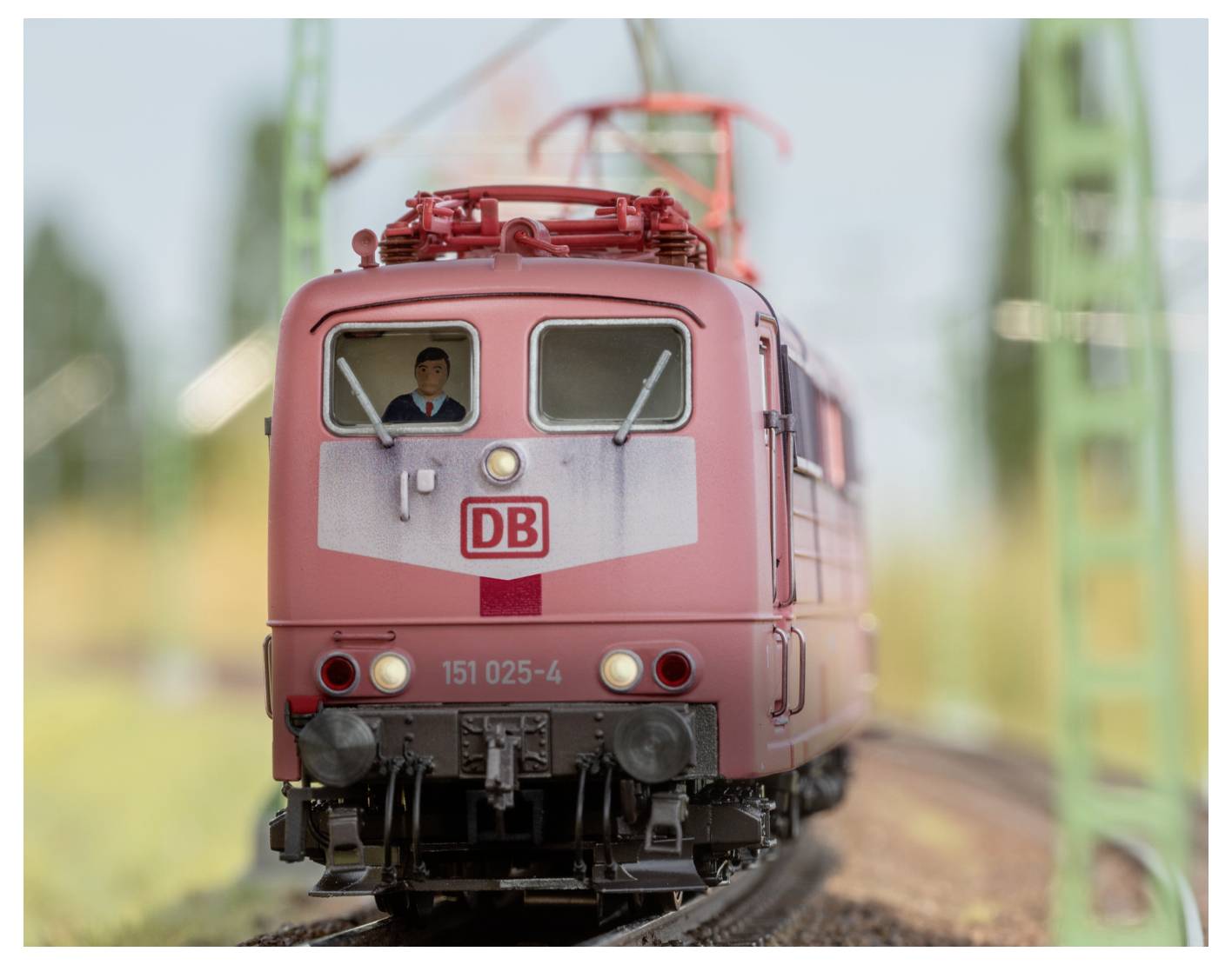 Egy piros vonat, amelynek homlokán a 'DB' logó és a '151 025-4' szám látható, vasúti síneken halad, elmosódott zöld háttérrel.