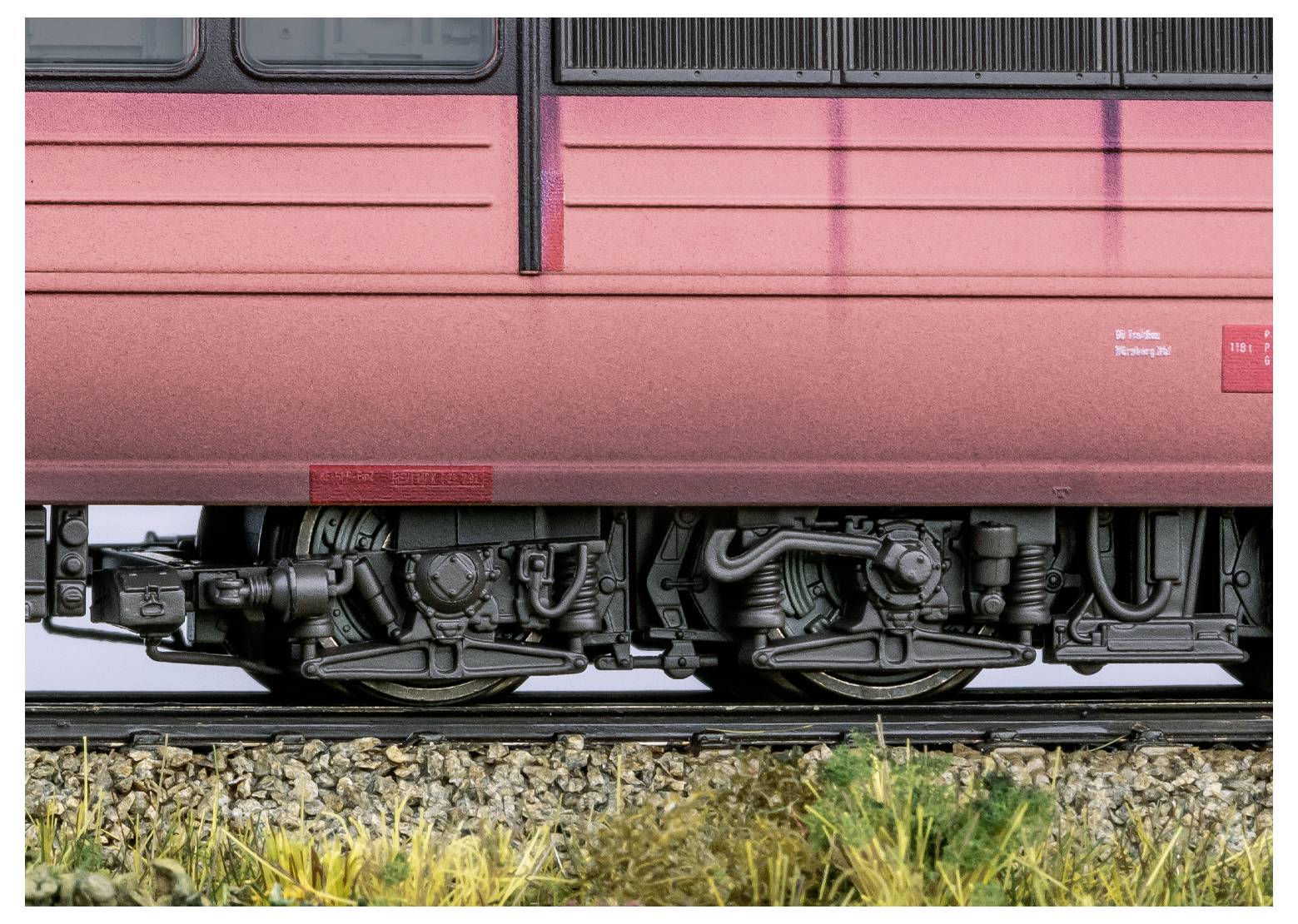 Vonatkerék közelképe a síneken, amely bemutatja az alváz aprólékos részleteit egy elmosódott vörös vonatháttér előtt.