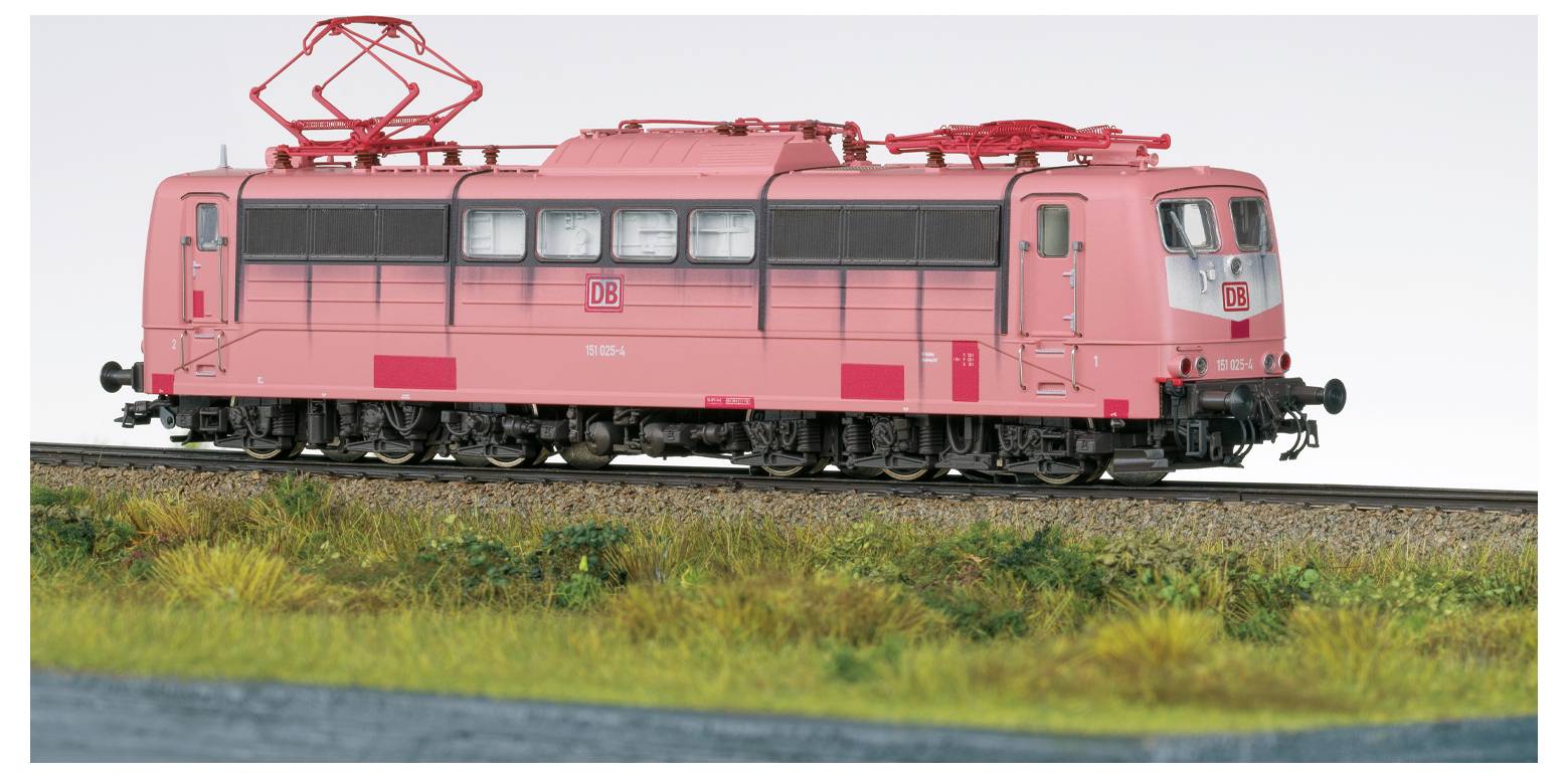 Egy DB logós élénkrózsaszín elektromos vonatmodell, amely síneken áll, fű veszi körül, tiszta égbolt alatt.