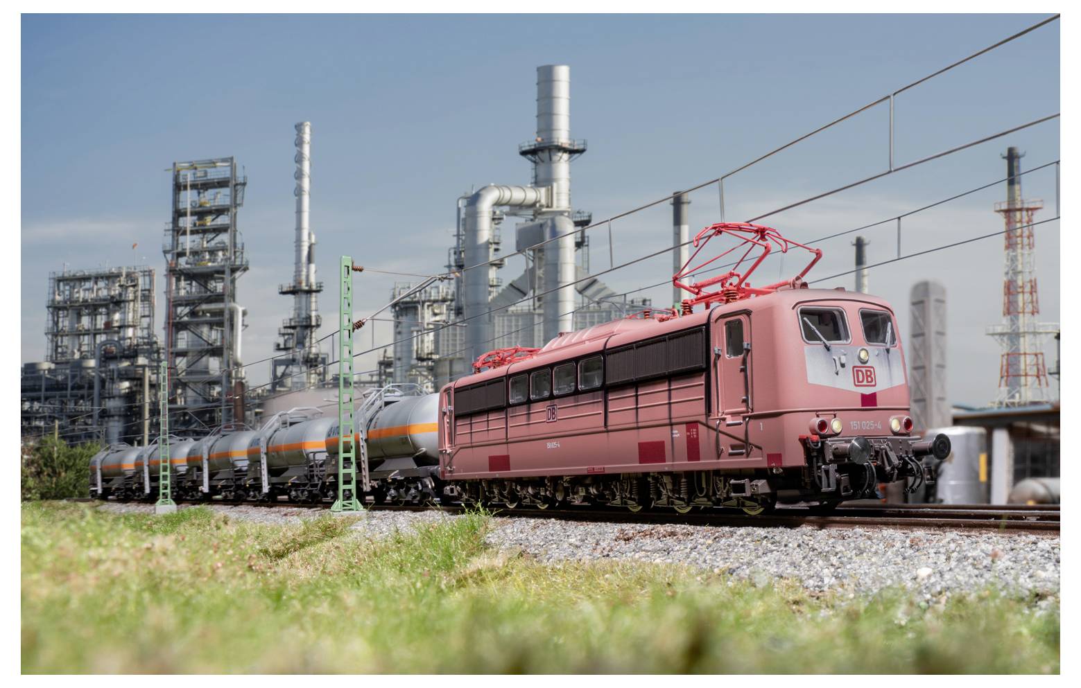 Egy piros vonat halad a síneken egy ipari üzem mellett, melynek háttérében magas kémények emelkednek egy kristálytiszta kék égbolt alatt.