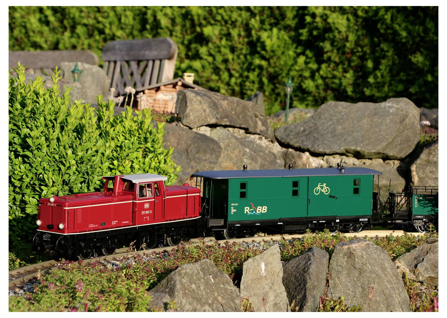 Egy modell piros mozdony húz egy zöld áruszállító kocsit, amelyen kerékpár szimbólum látható, amint áthalad egy kerti jelenetben, sziklák és növényzet között.