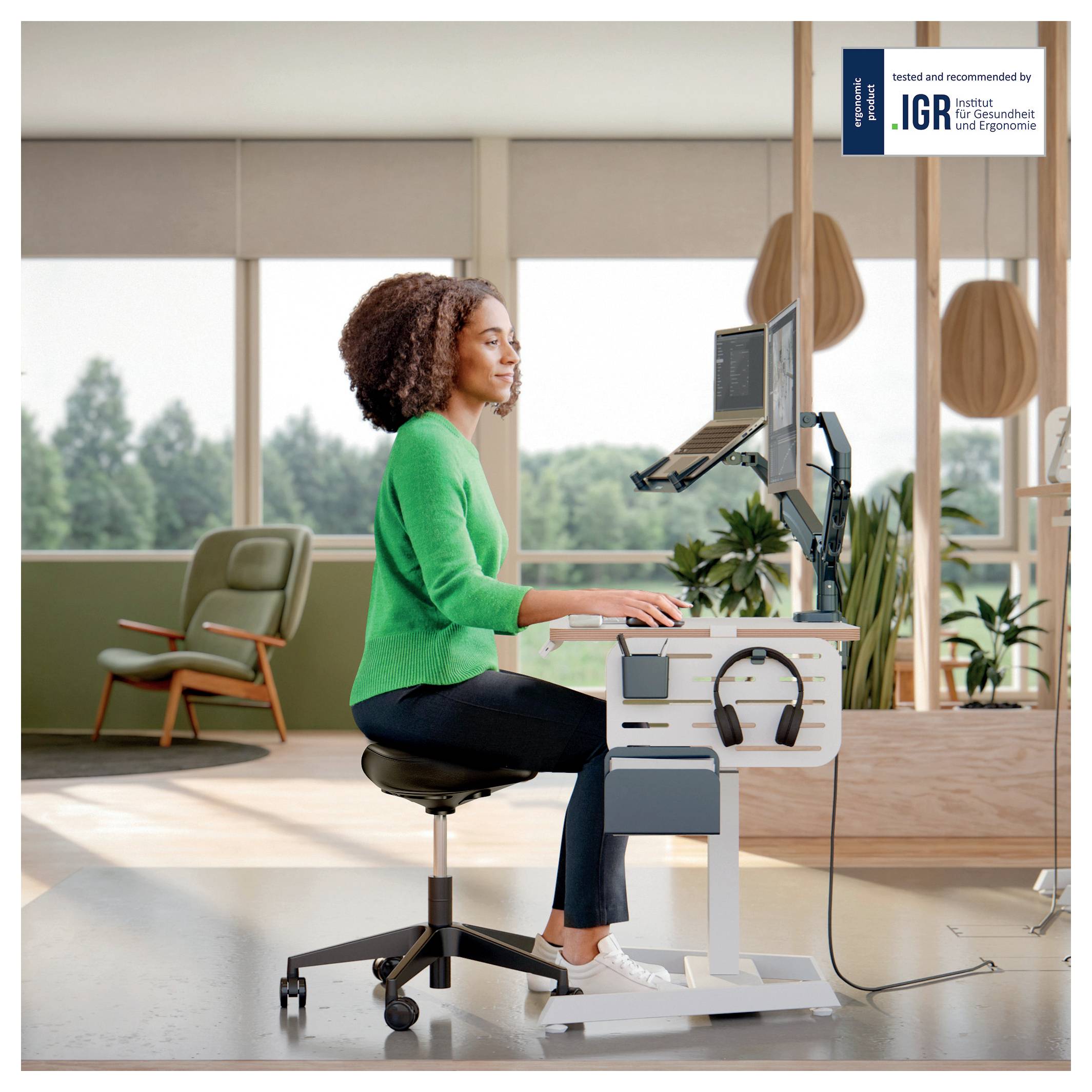 Egy nő egy ülő-álló íróasztalnál dolgozik egy modern irodában, természetes fénnyel körülvéve, egy ergonomikus székben ülve, számítógépet és tabletet használva.