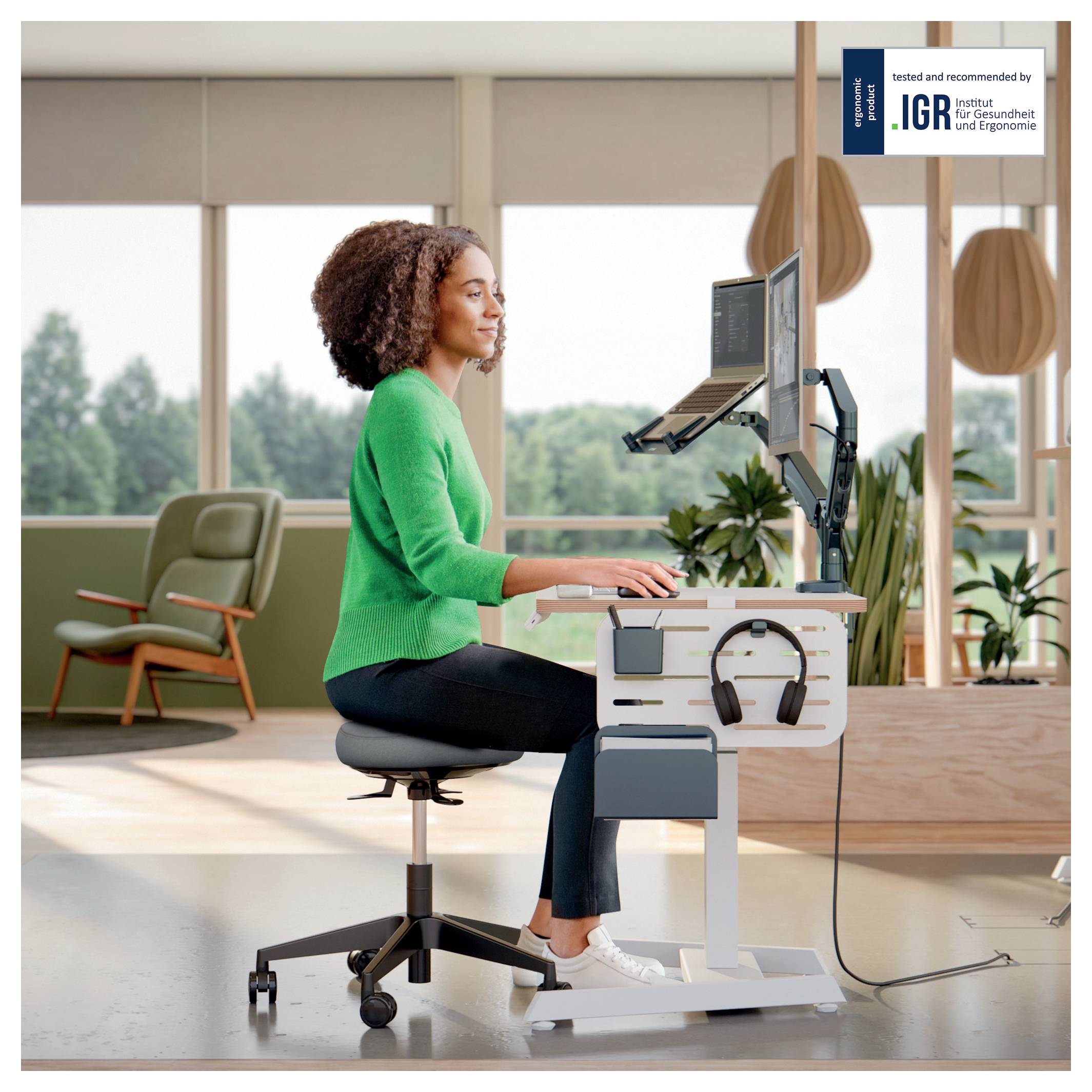 Egy személy ergonomikusan dolgozik egy állítható magasságú íróasztal mellett, miközben egy számítógépet használ. A háttérben nagy ablakok gondoskodnak a természetes fényről.