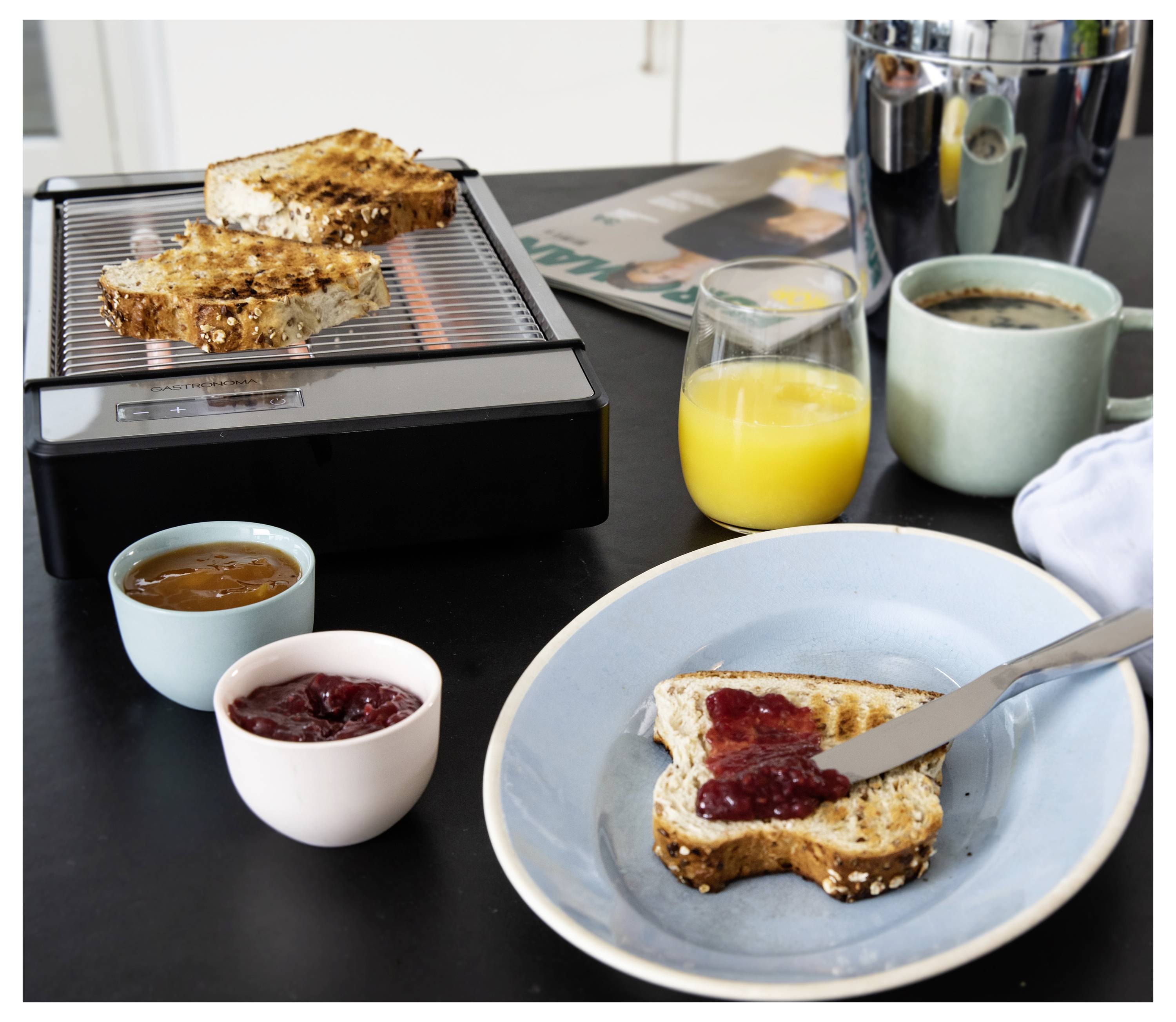 Reggeli jelenet egy tányéron két megpirult teljes kiőrlésű kenyérszelettel, amelyeket lekvárral kentek meg, mellette lekvár és narancsié egy asztalkán.