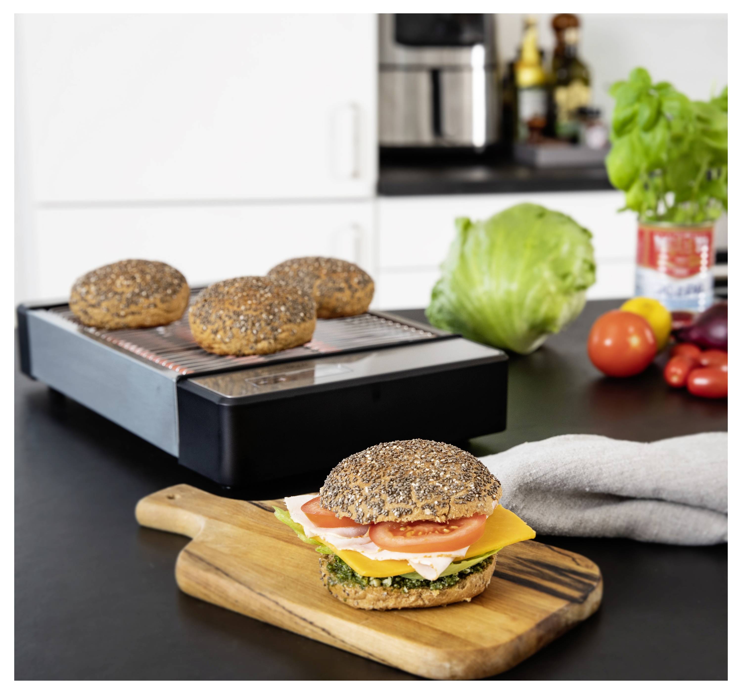 Egy szendvics sajt, paradicsom és saláta egy fadeszkán egy konyhában. Teljes kiőrlésű zsemlék vannak egy elektromos grillen a háttérben.