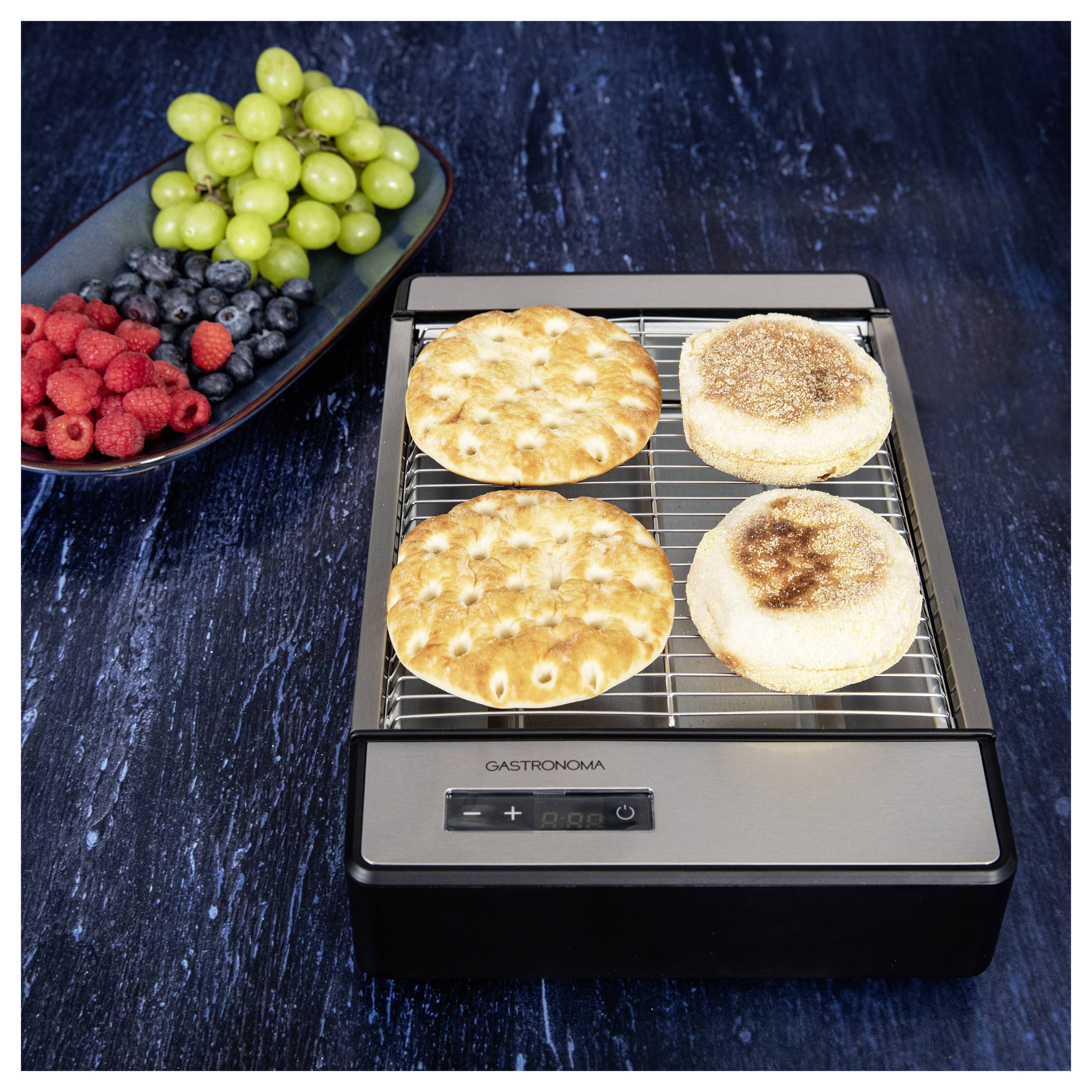 Két pitakenyér és két english muffin melegszik egy elektromos kenyérpirítón. Egy tányér szőlő, málna és áfonya van mellette.