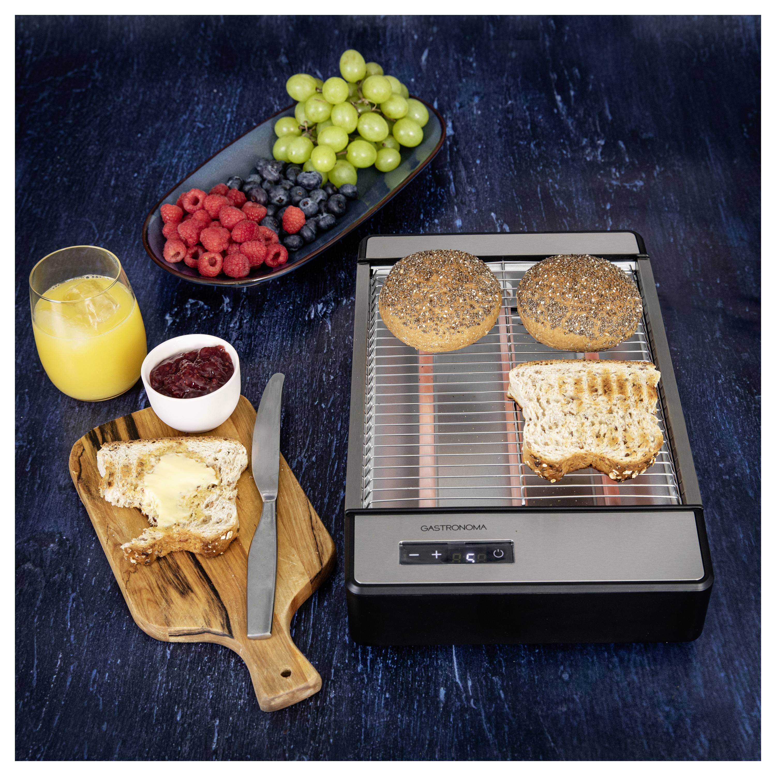 Reggeli terítés kenyérpirítóval, megpirított bagellel, vajazott kenyérrel, késsel, lekvárral, friss bogyókkal, szőlővel és egy pohár narancslével.