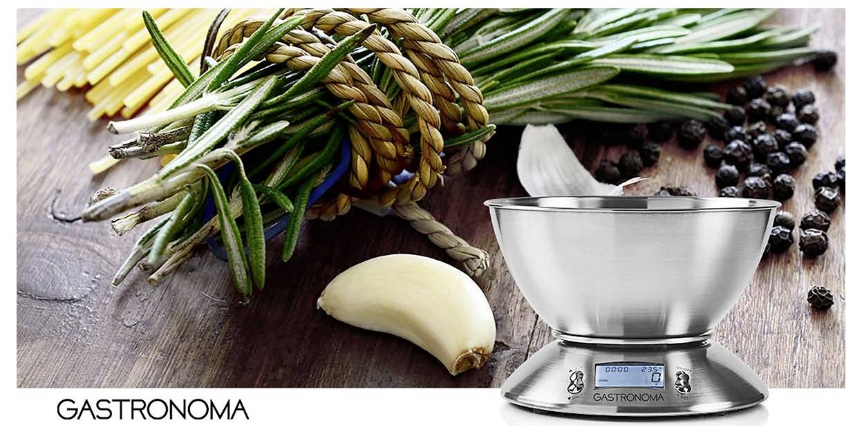 Egy fémes konyhai mérleg fa felületen, amelyen gyógynövények, fokhagyma és szemes bors látható. A 'Gastronoma' márkanév feltüntetve.