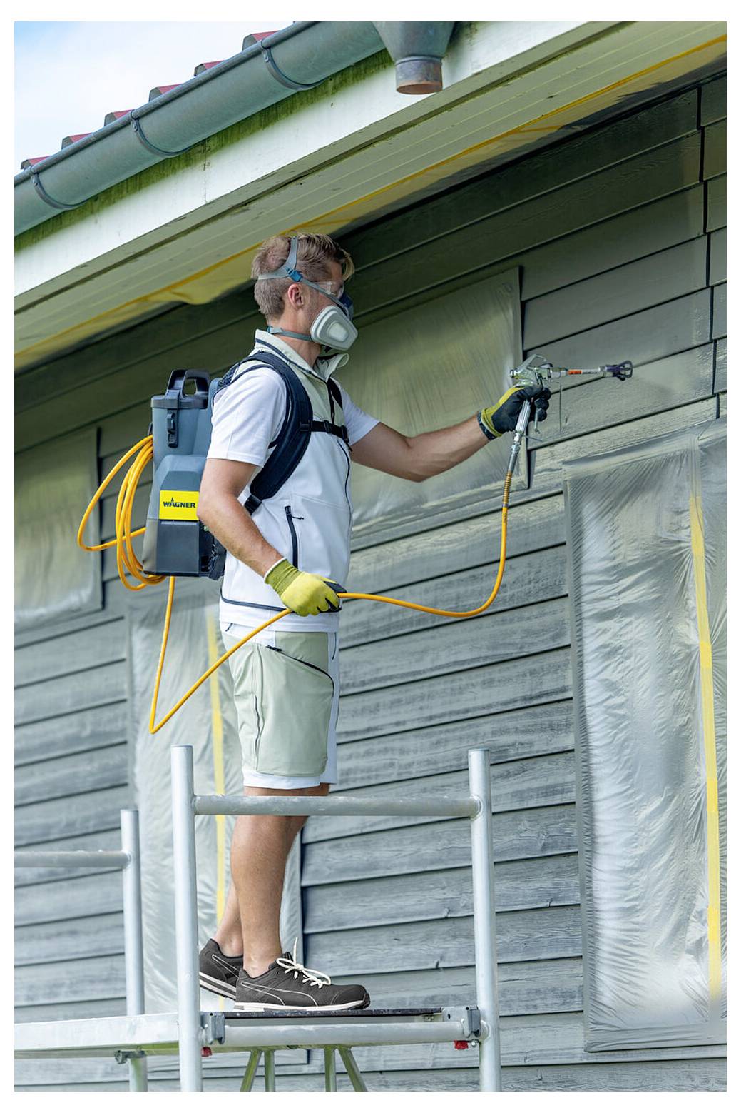 Egy védőfelszerelést viselő személy festékszórót használ egy ház oldalának festéséhez, állványzaton állva. A terület le van takarva védelemképpen.