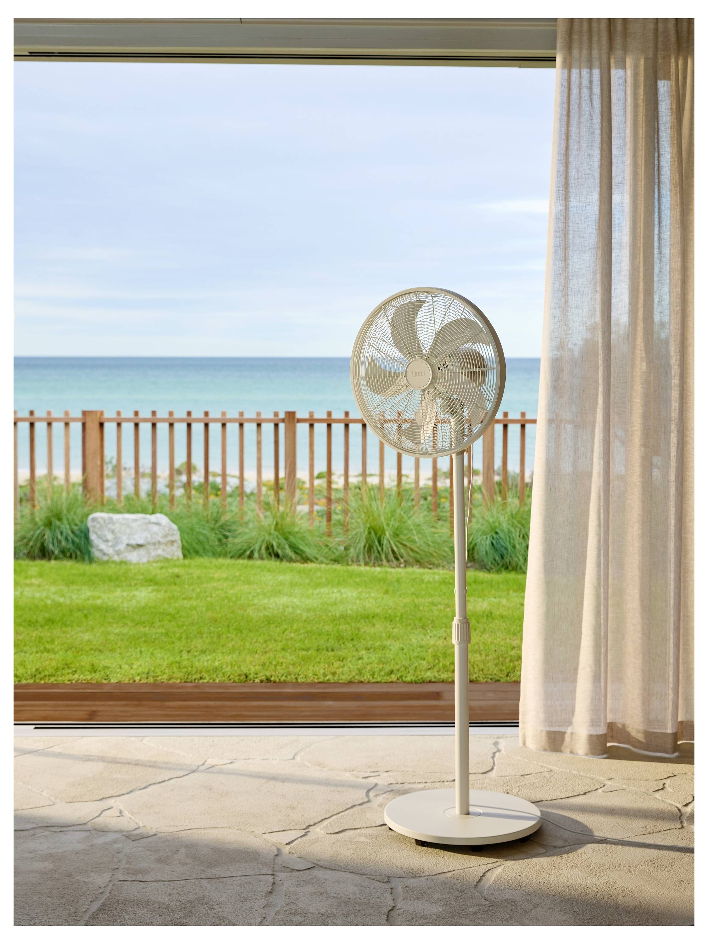 Egy álló ventilátor áll egy szobában, és egy nyitott tolóajtó felé néz, amelyen át egy fa kerítés, zöld fű és egy nyugodt óceán látható a felhős ég alatt.