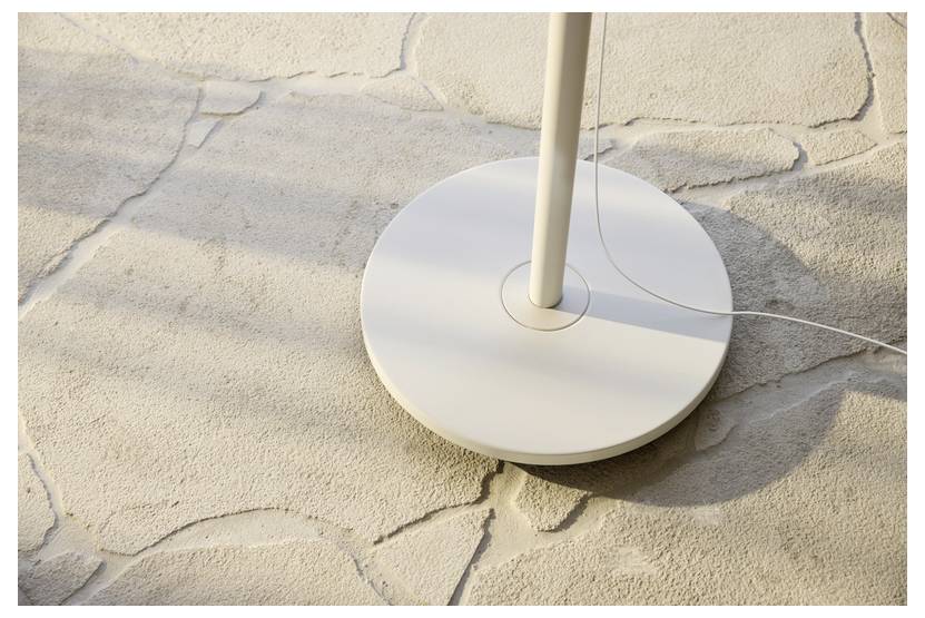 Egy fehér, kerek lámpatest csatlakoztatott zsinórral egy texturált kőpadlón ül, amelyen a napfény puha árnyékokat vet a felületre.
