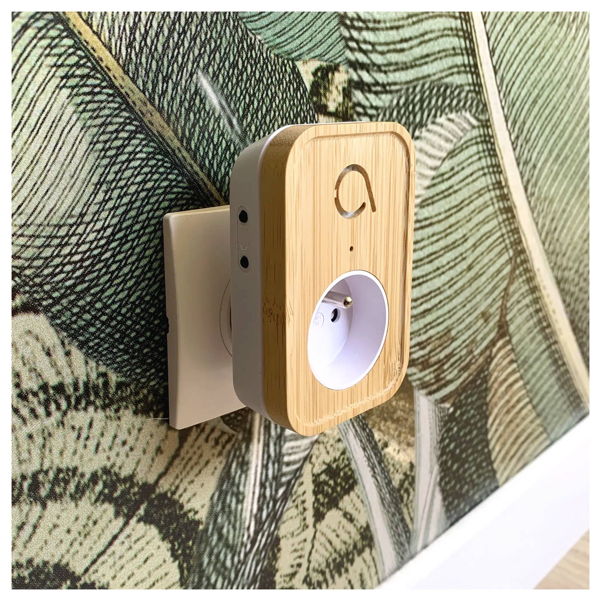 Egy okos dugalj bambusz felülettel be van dugva egy fali konnektorba, zöld és fehér mintás tapéta előtt.