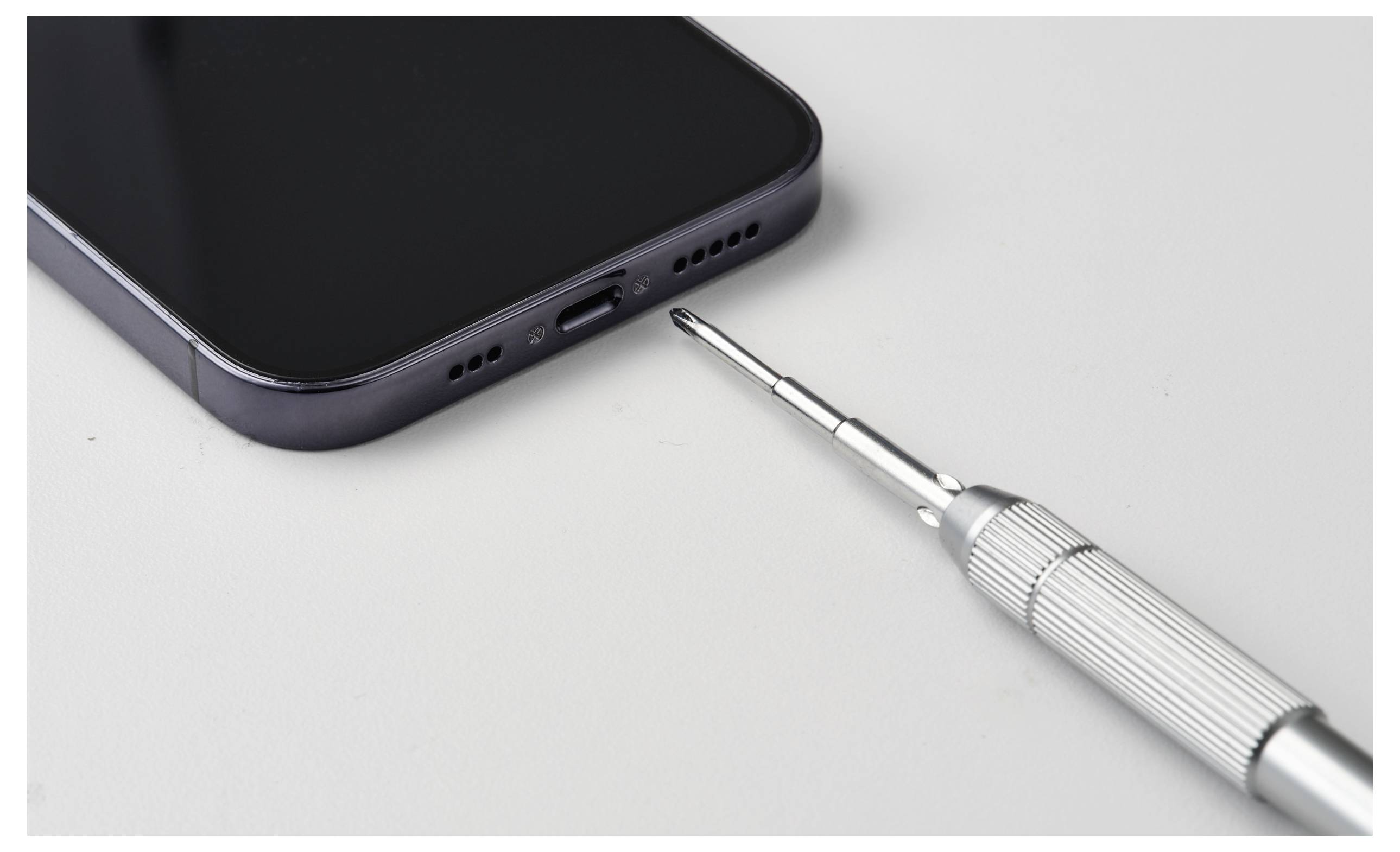 Egy okostelefon fekete képernyővel és egy precíziós csavarhúzó mellette fehér felületen, amely telefonjavítást vagy karbantartást jelez.